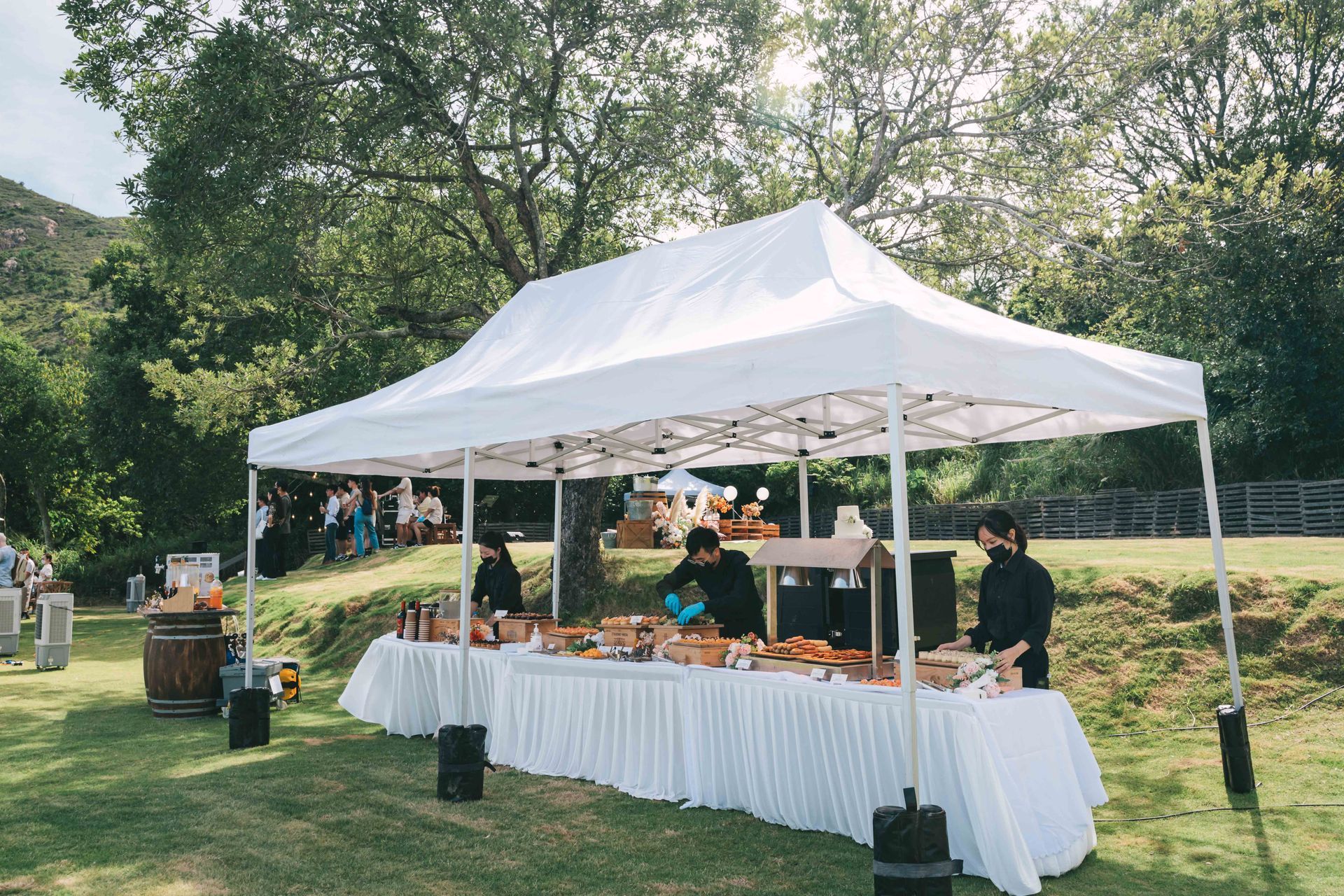 Food serving station set up under a white tent in a park. Servers behind a long table with food.