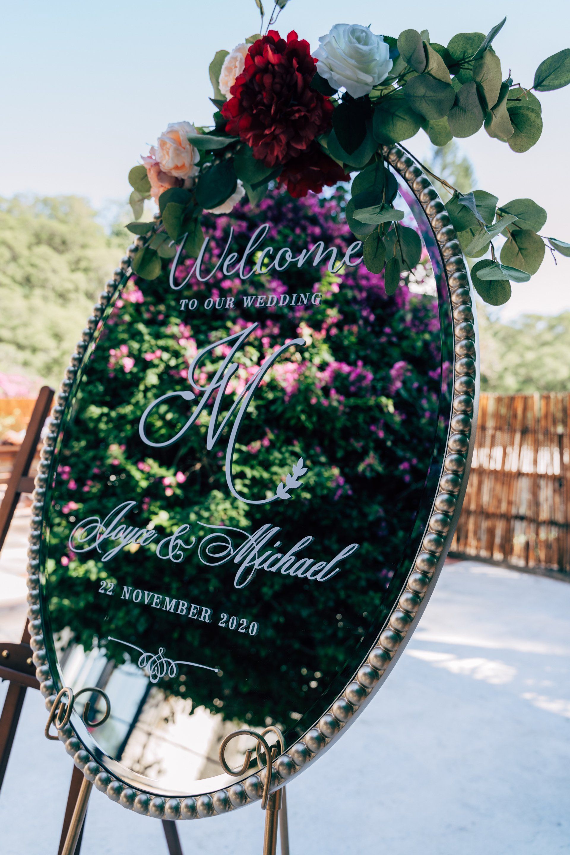 Wedding welcome sign on a decorative mirror with floral accents and names: Anya & Michael, displayed outdoors.