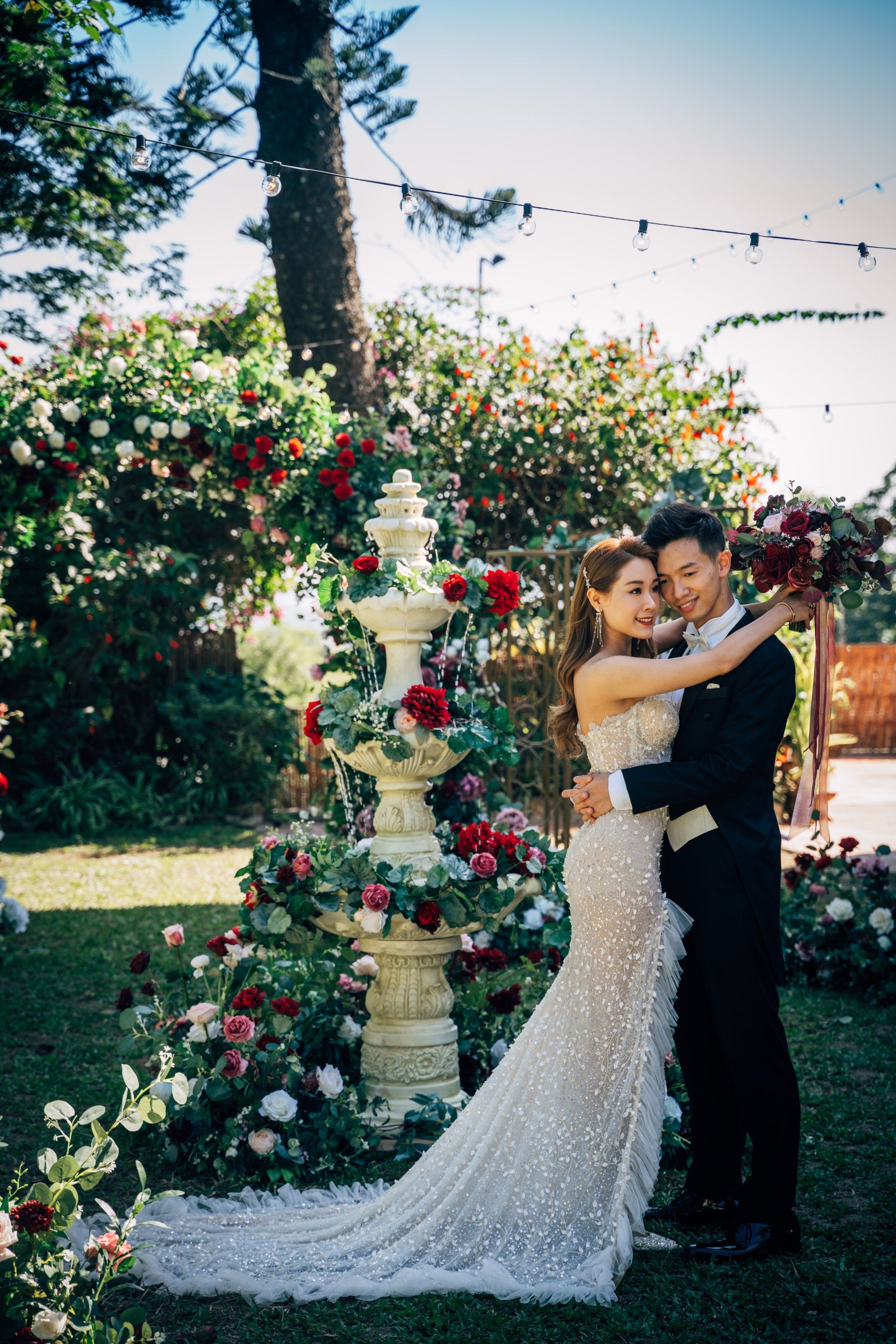 Couple embracing in a garden, near a decorated fountain. Bride in a beaded gown, groom in a tuxedo. Lush flowers.