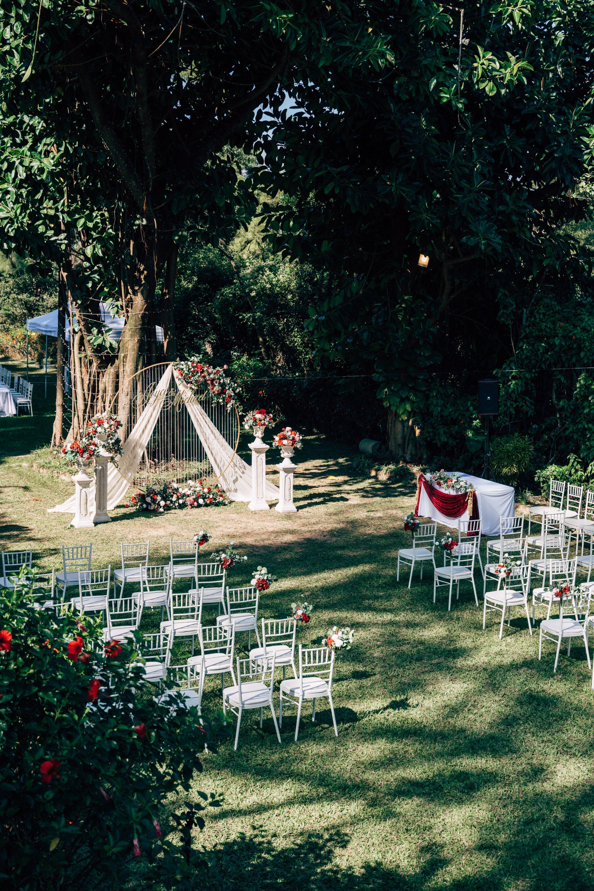 Wedding ceremony setup in a sunny garden: white chairs, flower arrangements, arch, trees.