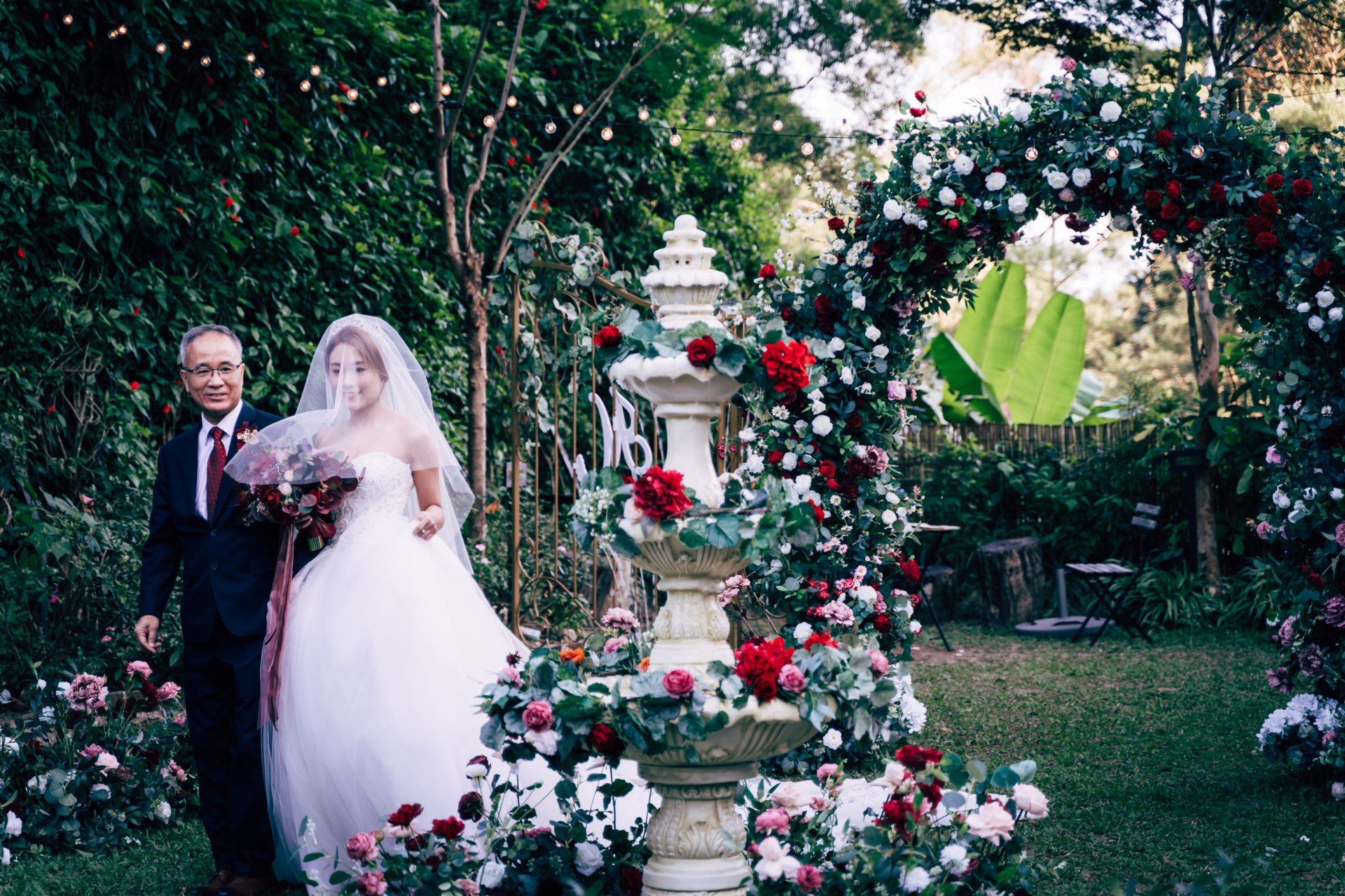 Bride in white dress walks down an aisle with her father, passing a floral arch and fountain.