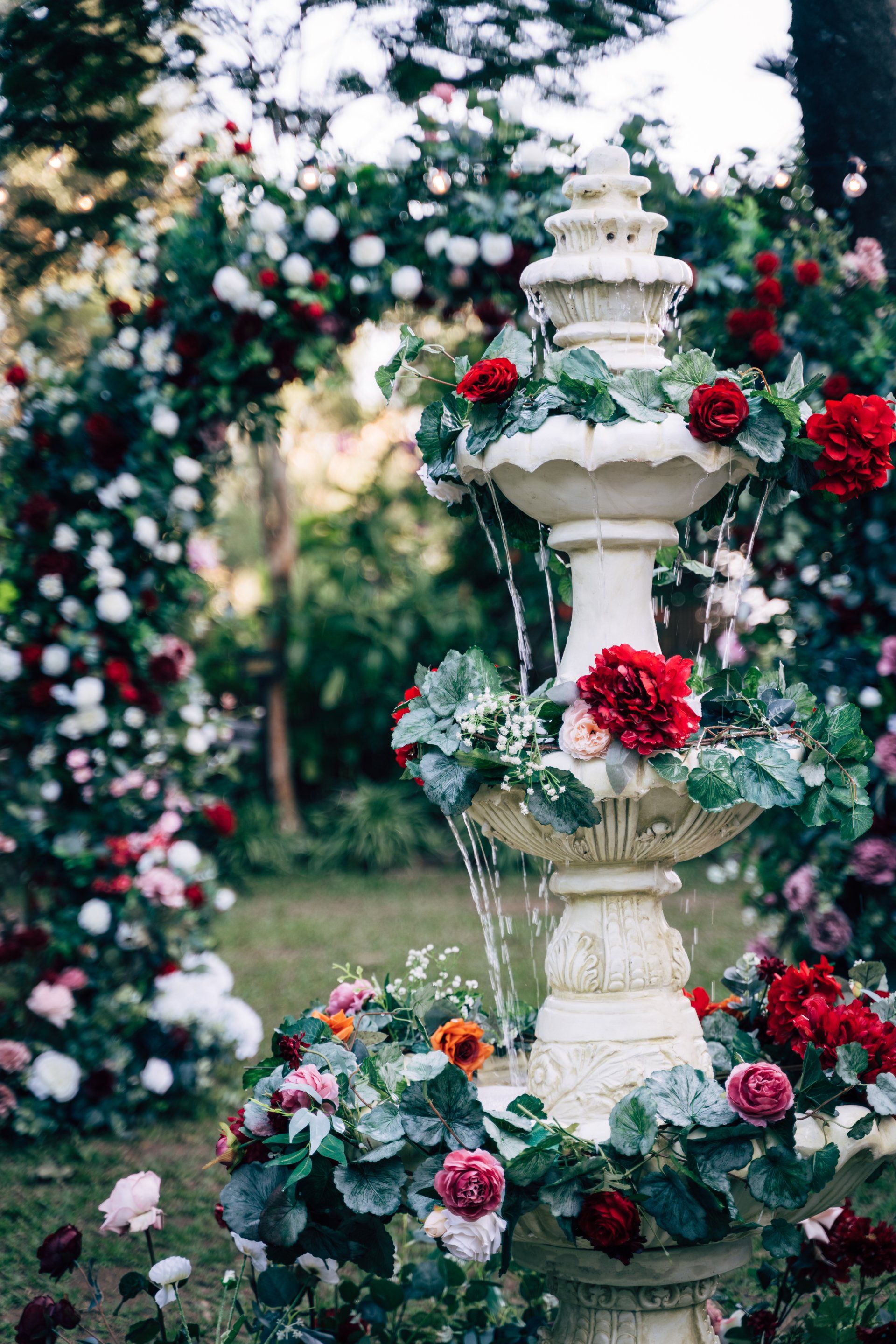 Ornamental fountain with cascading water, adorned with red and pink flowers, in a garden setting.