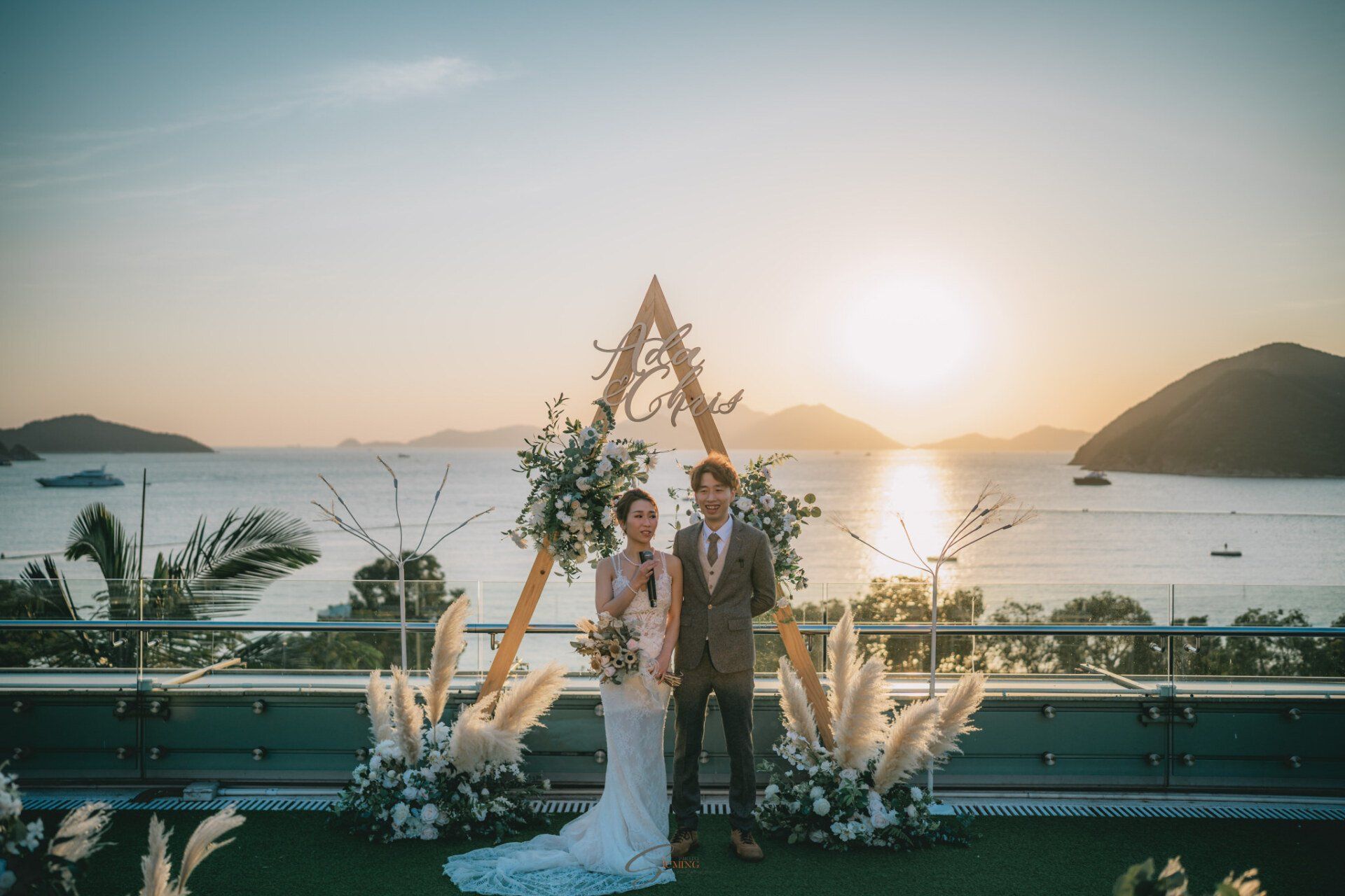 Couple stands before a floral arch on a rooftop overlooking a bay at sunset.