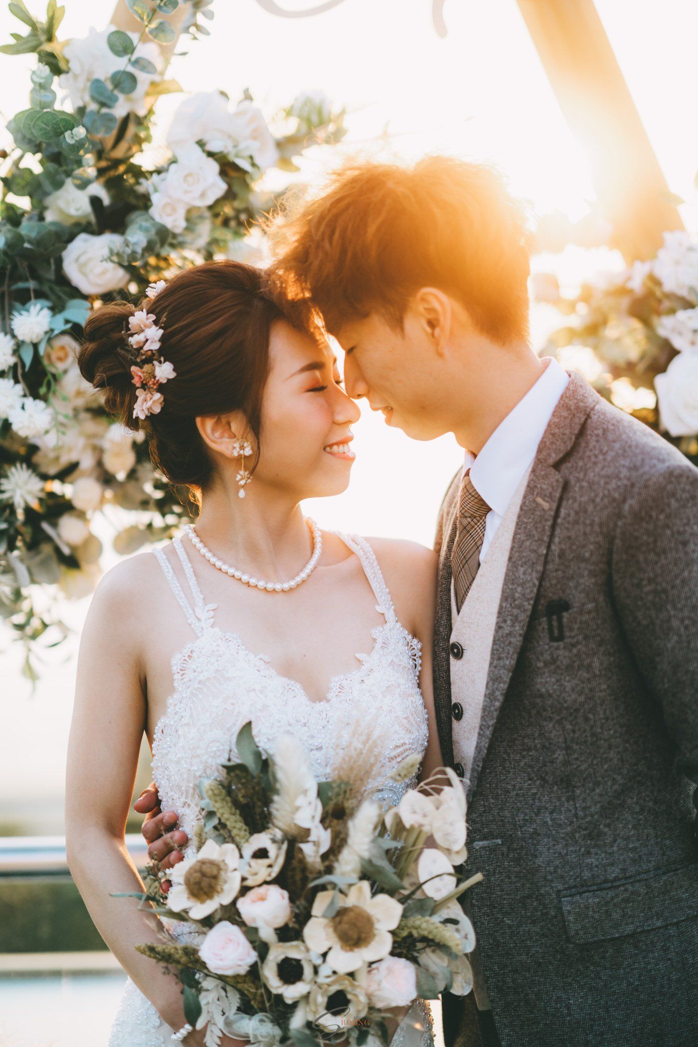 Couple embracing, wedding ceremony. Bride in white dress, groom in gray suit. Sunlit arch with flowers.