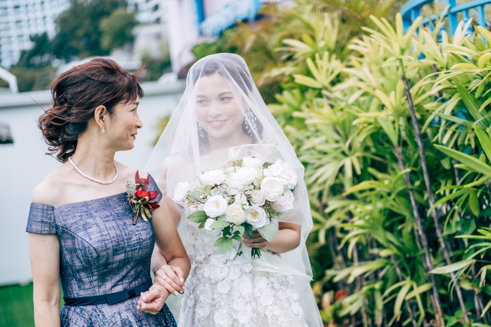 Bride in veil with bouquet, walks with woman in blue dress outdoors.