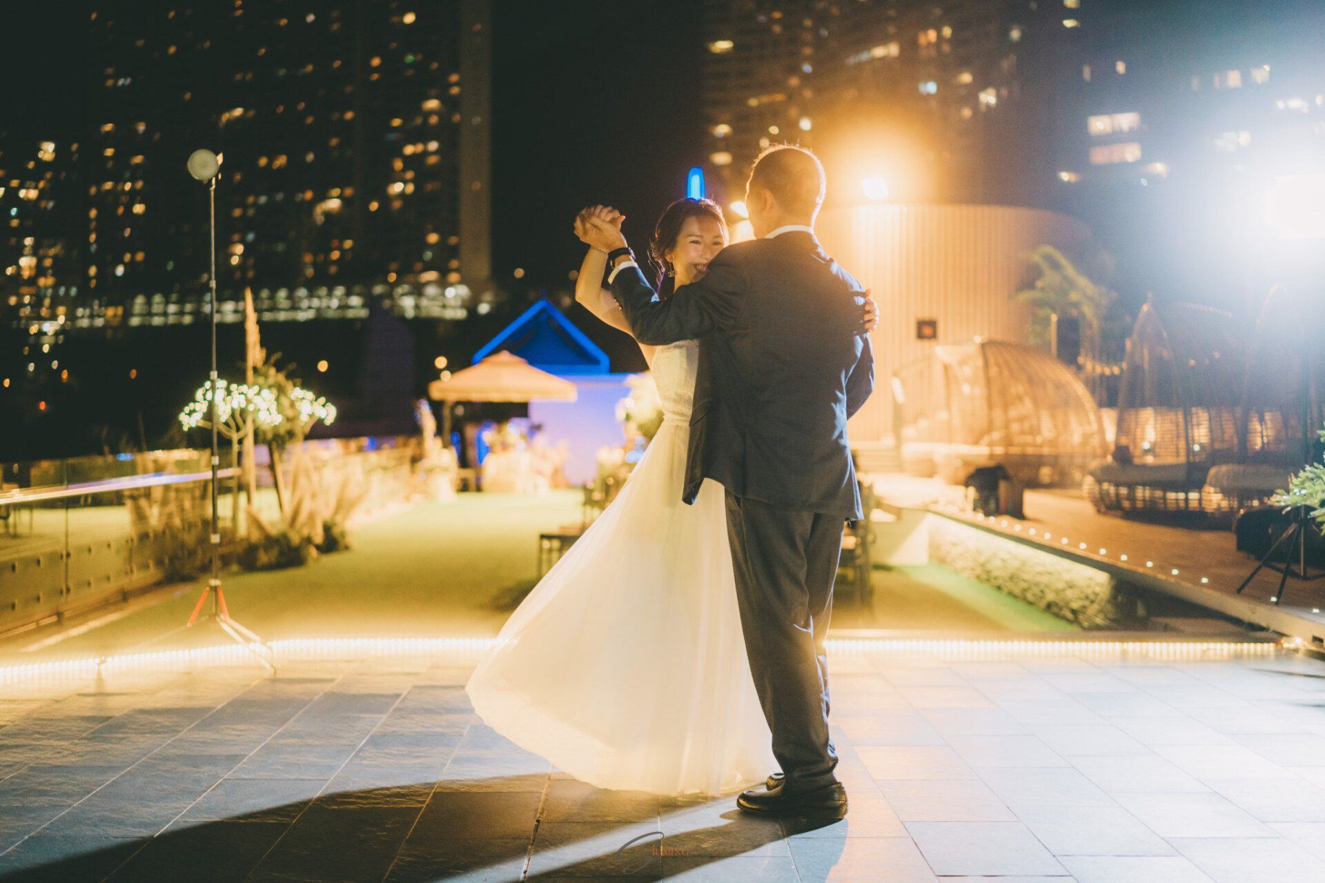 Couple dancing at a nighttime rooftop wedding reception; city lights in the background.