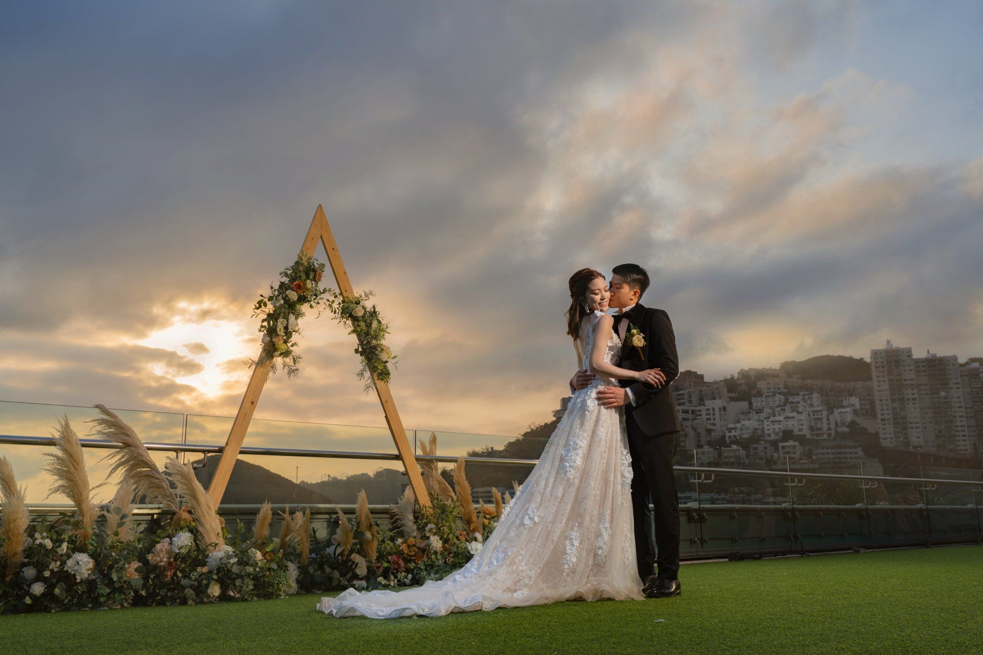 Couple embraces on a rooftop at sunset, floral arch in the background, city view.