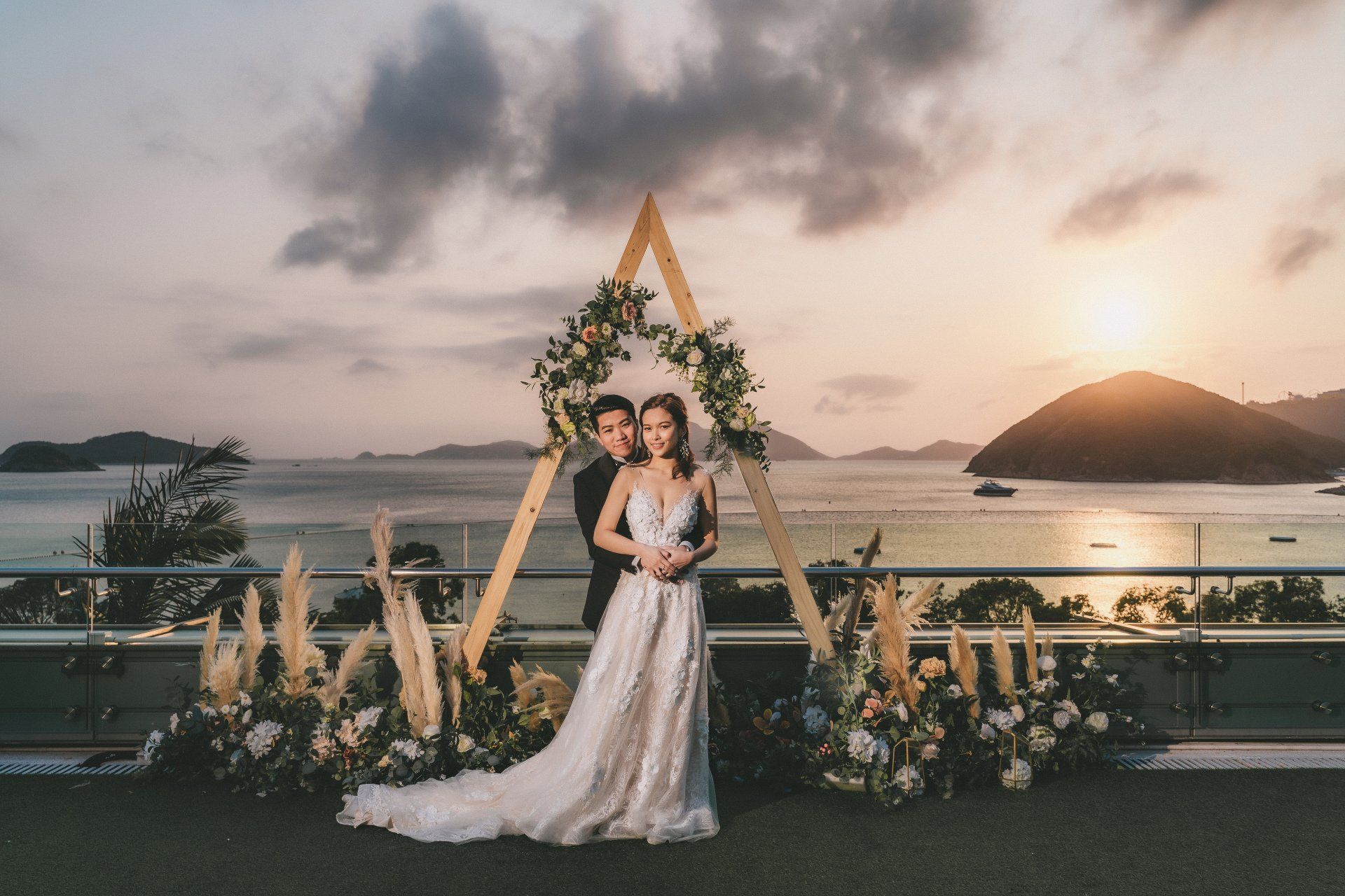 Couple embraces under a wooden arch decorated with flowers, overlooking ocean at sunset.