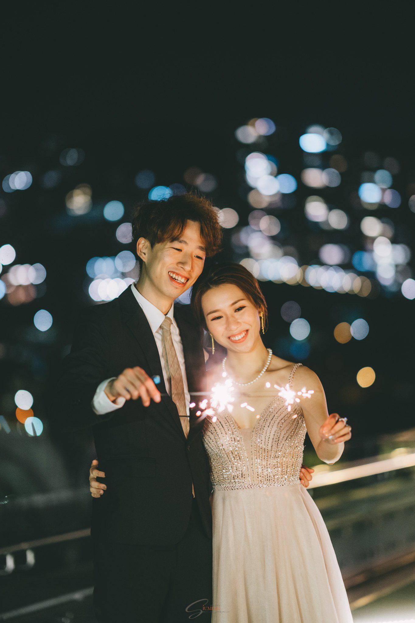 Couple smiles, holding sparklers at night, city lights blurred in the background.