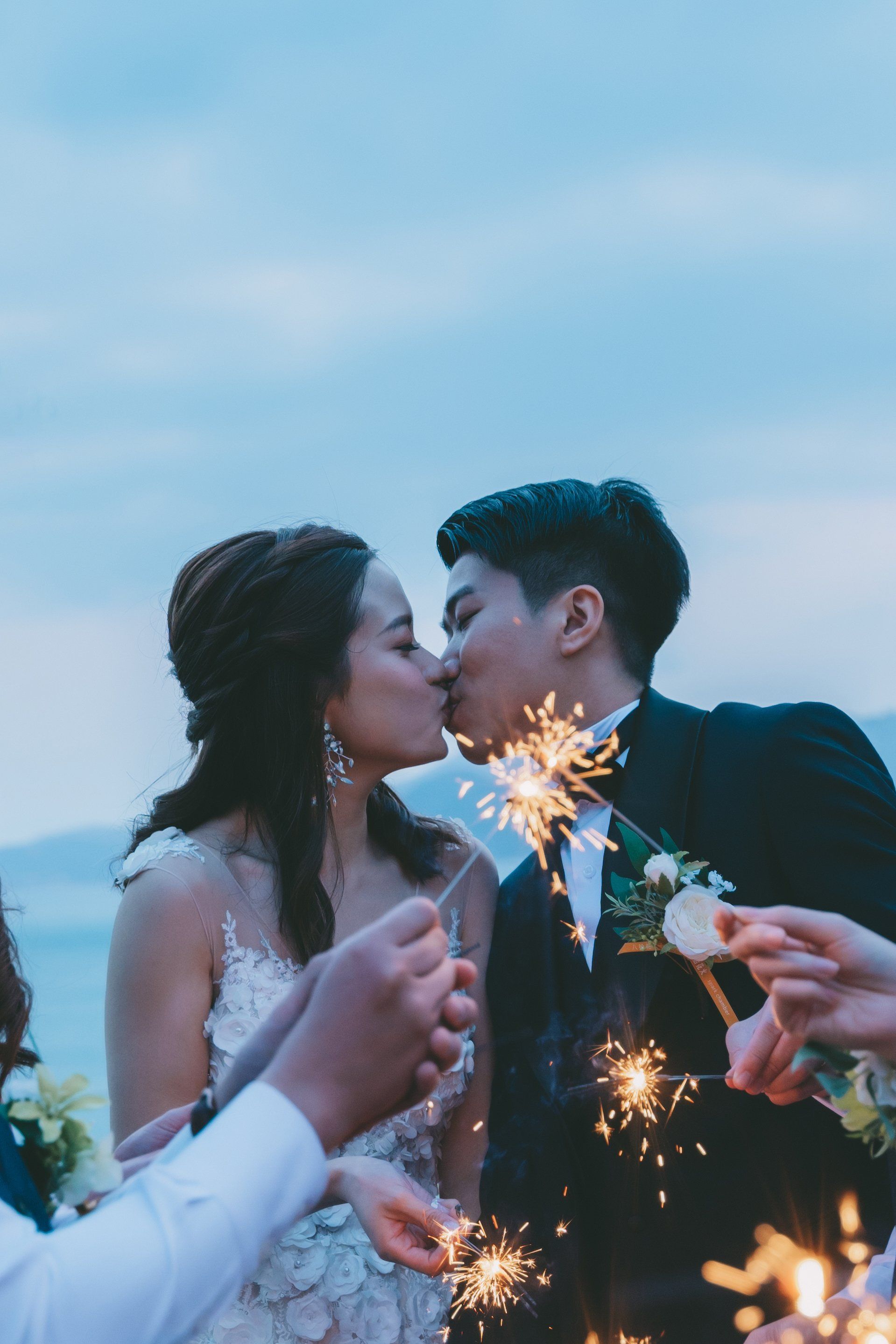 Couple kissing, holding sparklers at a wedding, celebrating with friends.