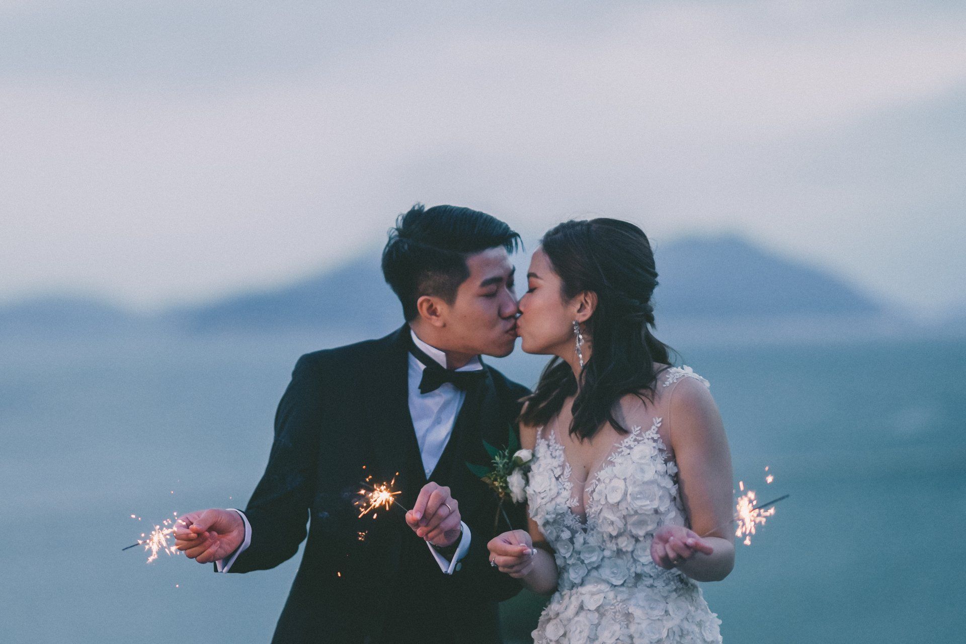 Couple kissing, holding sparklers at a wedding, with ocean and mountains in the background.