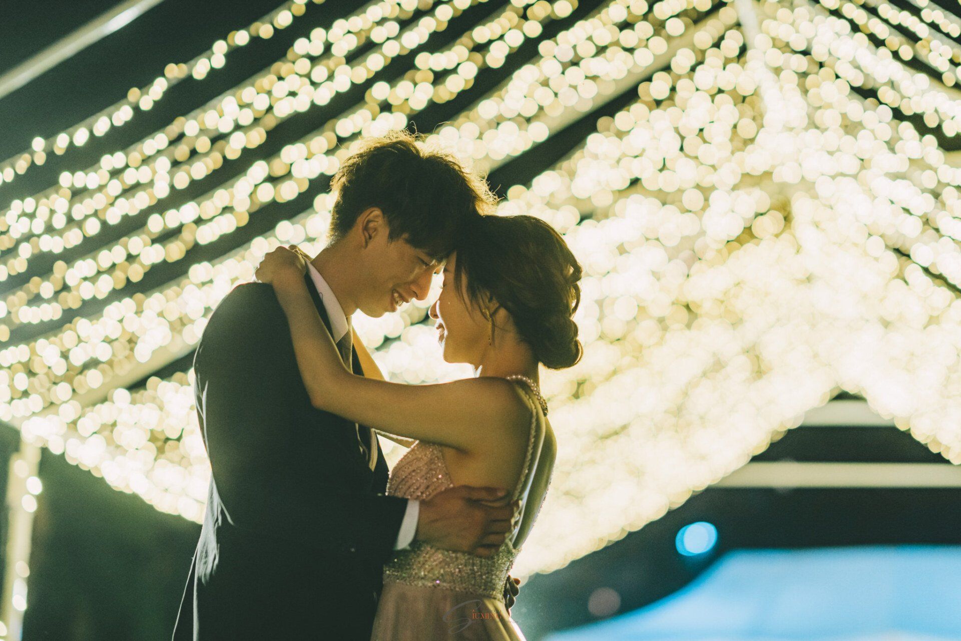 Couple embraces, illuminated by string lights, at an outdoor event.