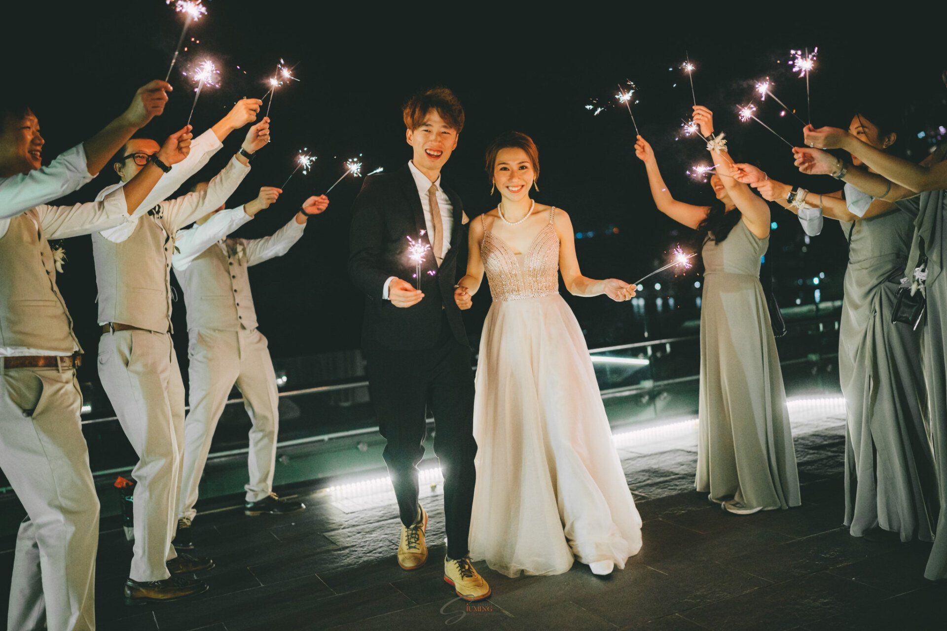 Bride and groom walk through sparkler-wielding wedding party at night; city lights in background.