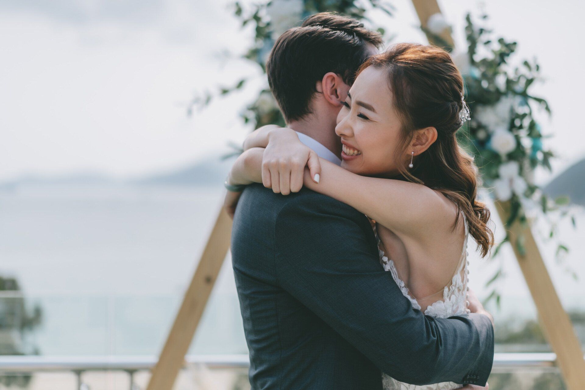 Bride hugging groom with ocean view; floral arch in background.