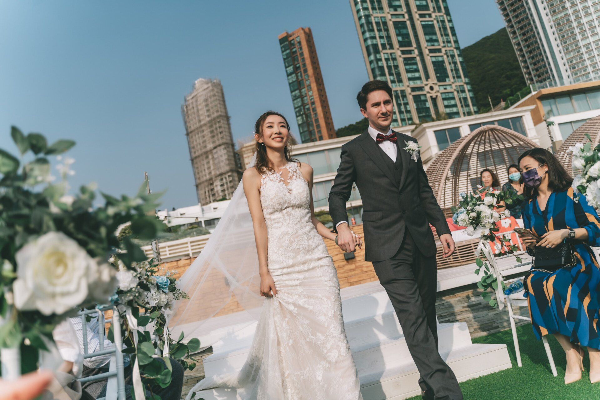Newlyweds walk hand-in-hand down an aisle after a wedding ceremony, against a backdrop of city buildings.