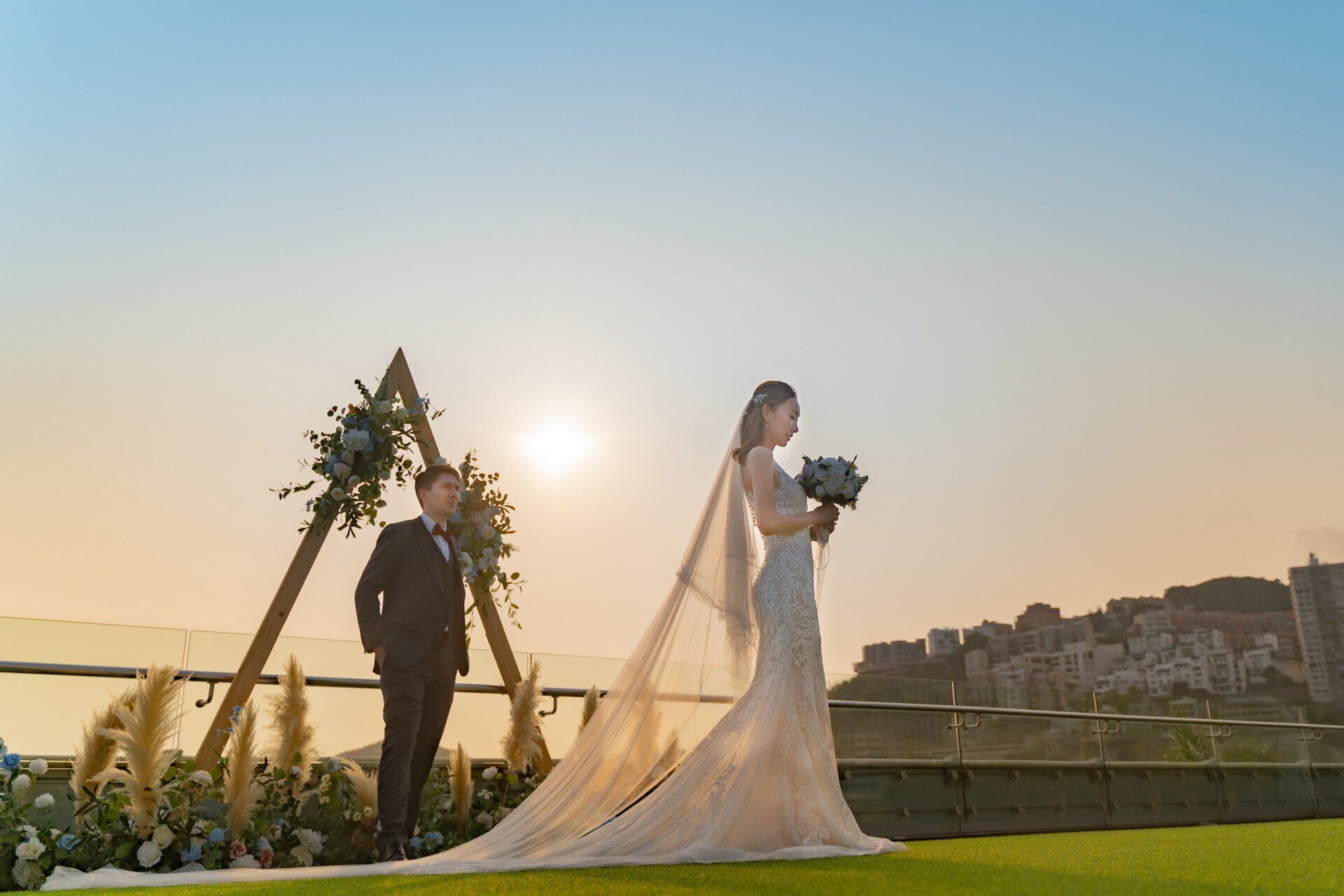Bride in a wedding gown holding a bouquet; groom stands before a decorated arch. Sunset backdrop.