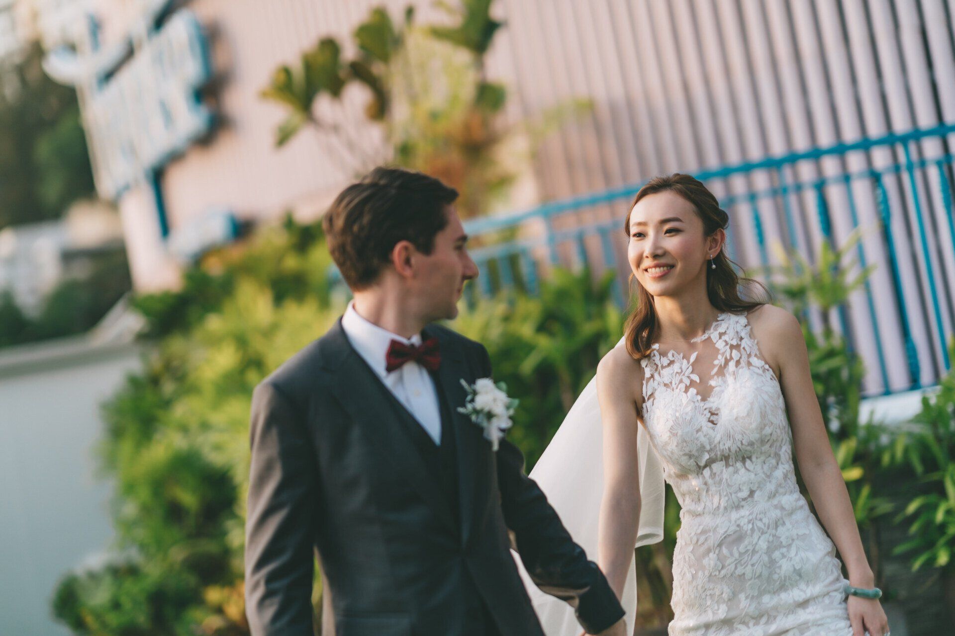 Bride and groom hold hands, smiling at each other. He wears a suit with bow tie. She wears a lace wedding dress.