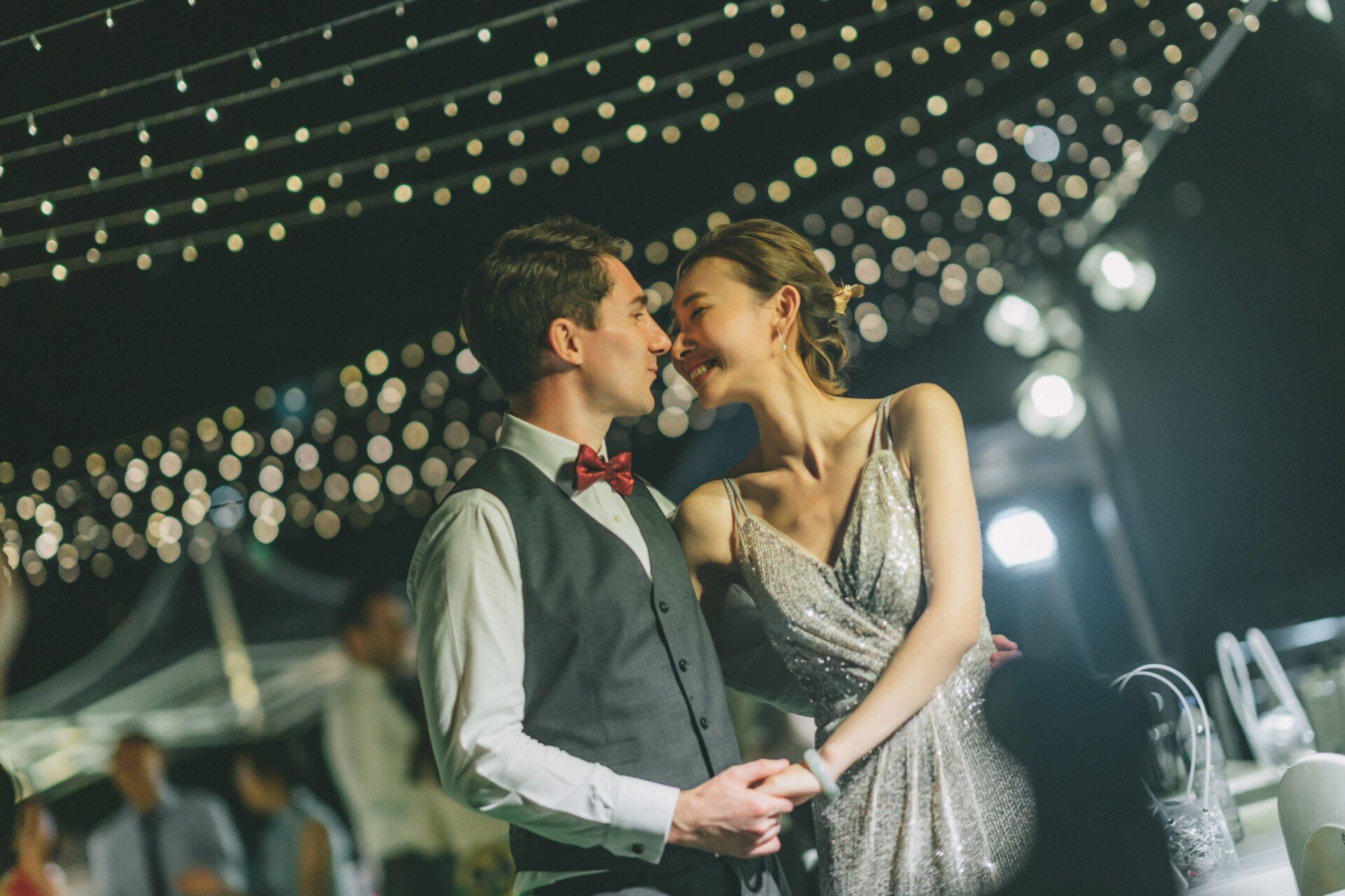 Couple smiles, almost kissing, under string lights. Man in vest and bow tie. Woman in sequined dress. Evening.