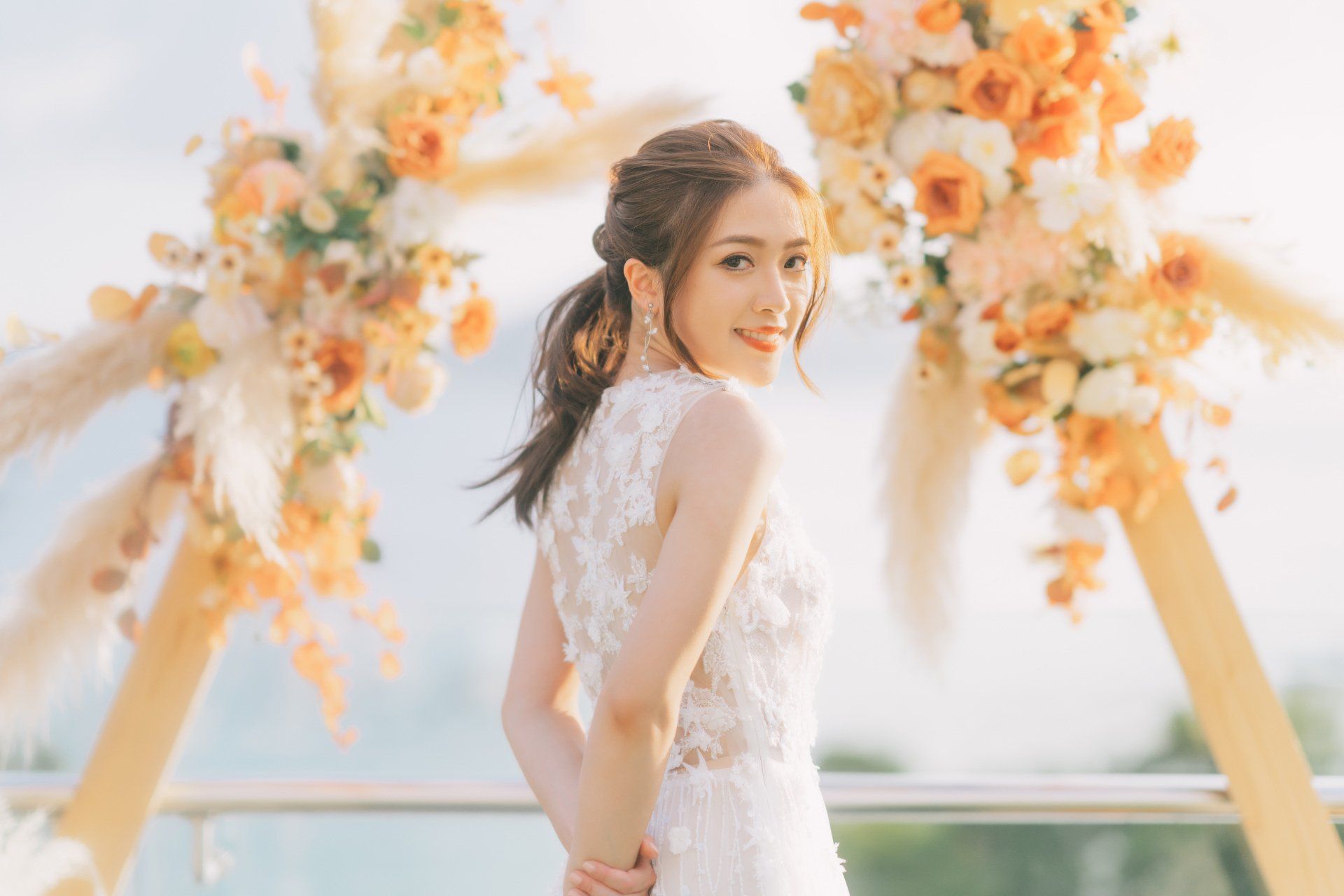 Woman in a white dress, smiling, near floral arch with orange and cream flowers; outdoor setting.
