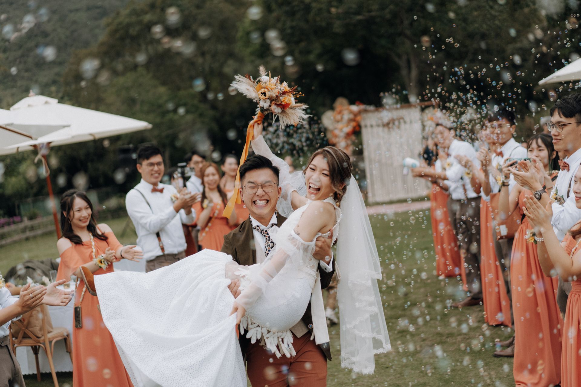 Groom carries bride at outdoor wedding, surrounded by cheering guests, bubbles, and orange-toned attire.