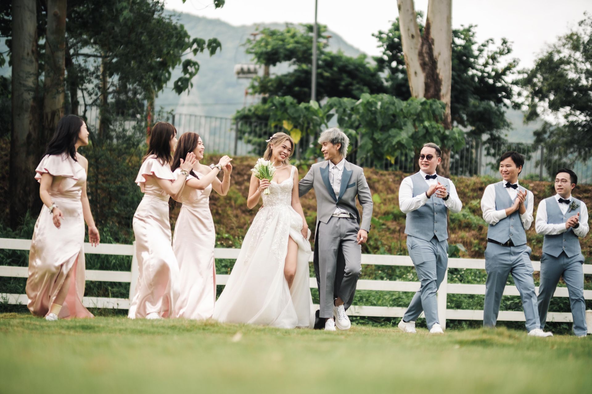 Wedding party: bride, groom, bridesmaids, and groomsmen walking on grass near a white fence.