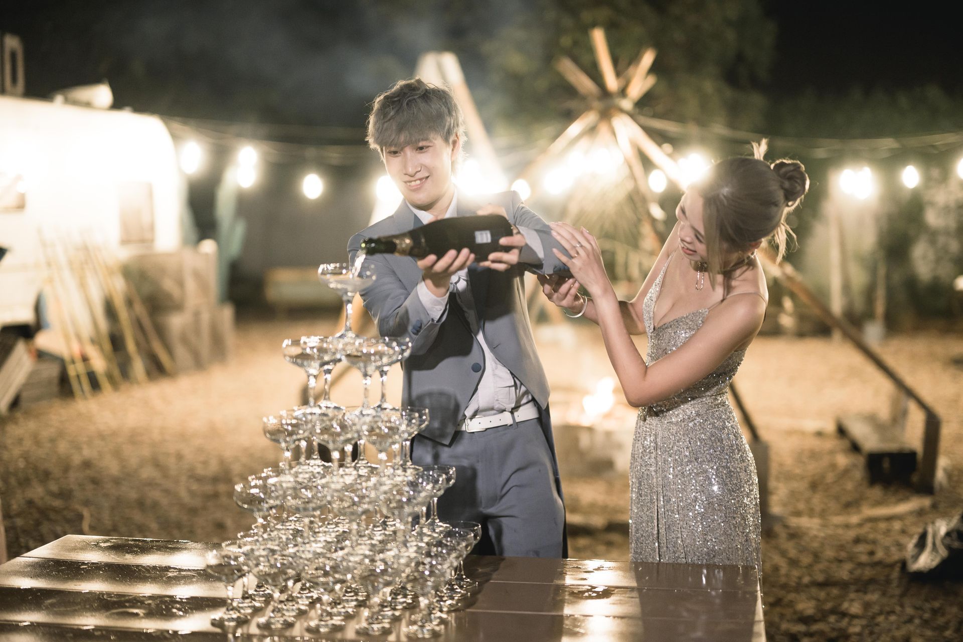 Man pours champagne into a tower of glasses, celebrating with woman. Outdoor evening event.