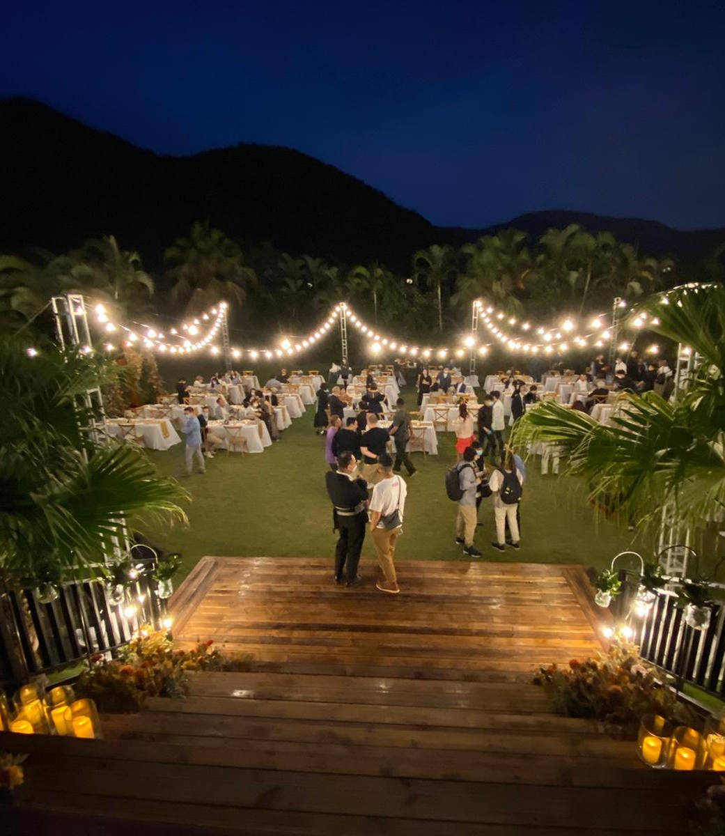 Outdoor evening event with string lights, tables, and people on a green lawn.