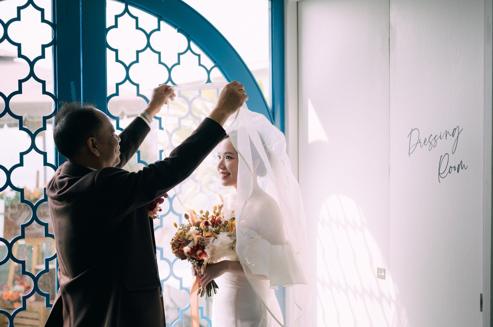 Father adjusting bride's veil. Bride holds bouquet; both stand near window. Sunlight streams in.