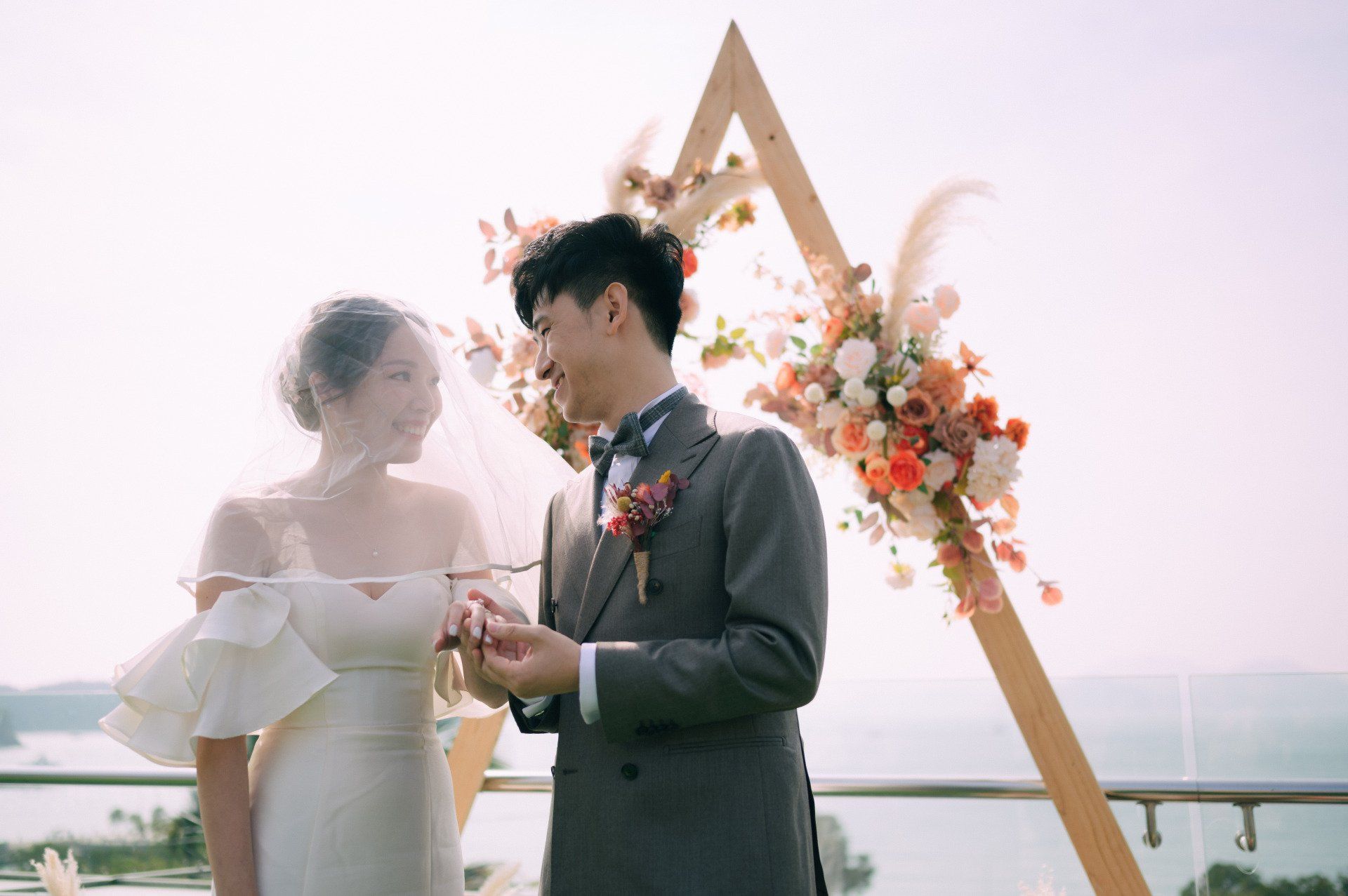 Bride and groom at outdoor wedding, smiling. Floral arch in background, water view.