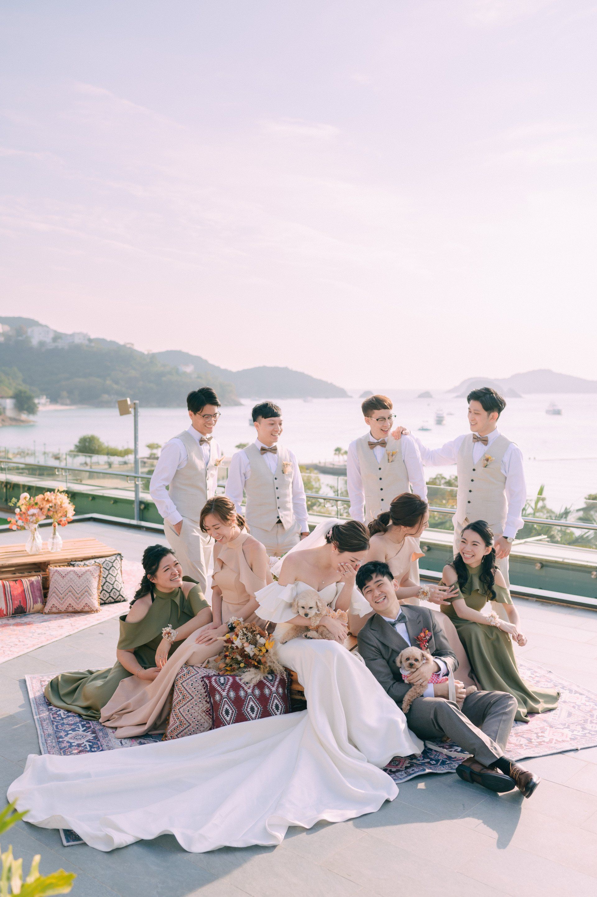 Wedding party posing on rooftop with ocean view. Bride in white dress, groom in grey, bridesmaids in pink/green, groomsmen in vests.