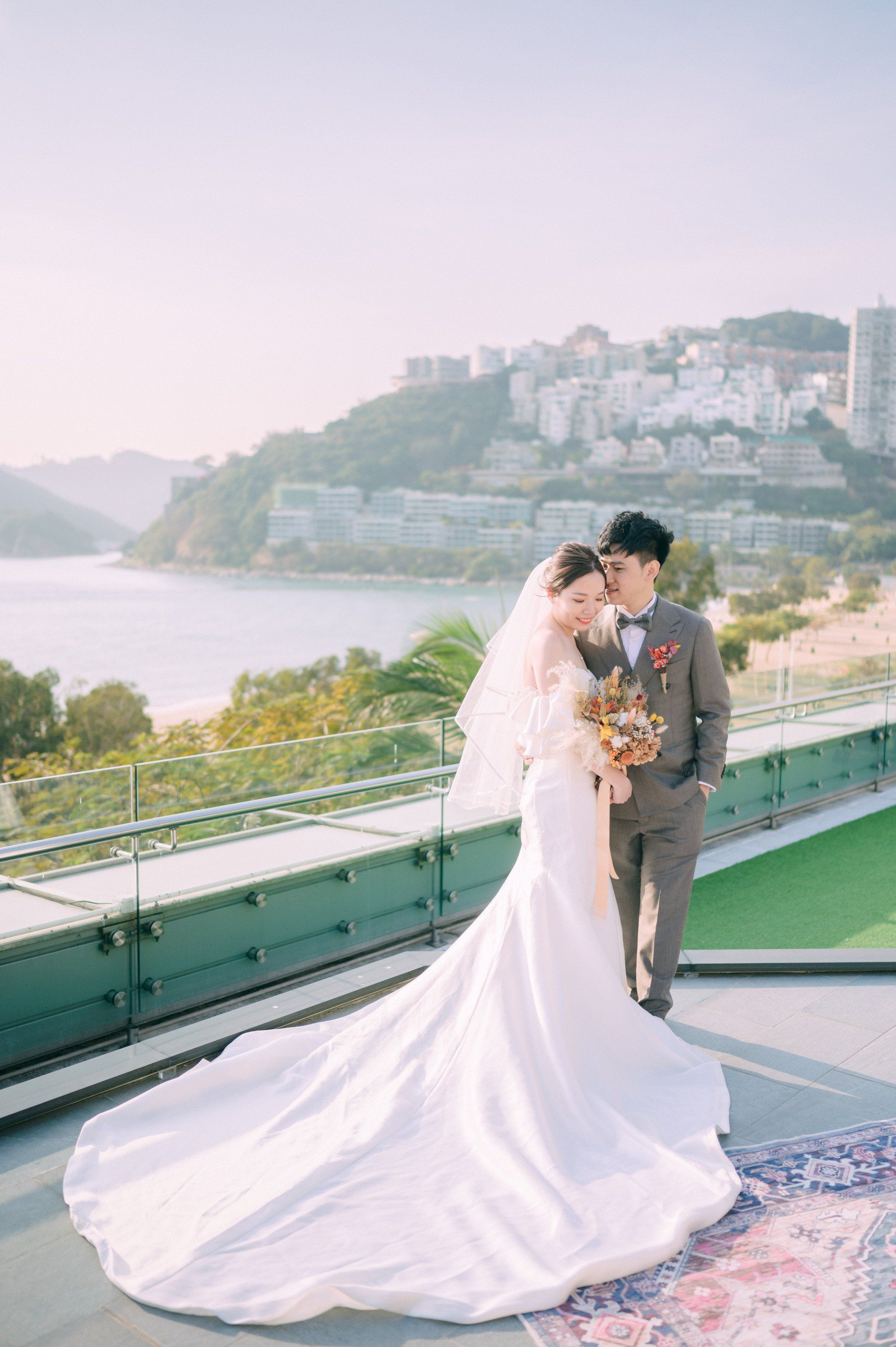 Bride and groom pose on a terrace overlooking a bay. The bride wears a white gown with a long train; the groom wears a gray suit.