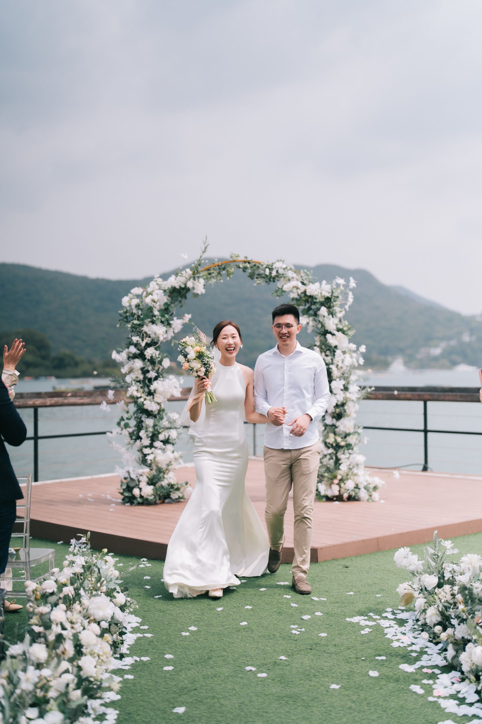 Newlyweds exiting wedding ceremony, holding hands, smiling. Floral arch with water and mountains in the background.