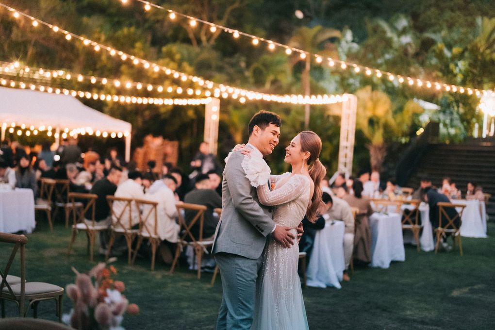 Couple dancing at outdoor wedding reception with string lights and guests.