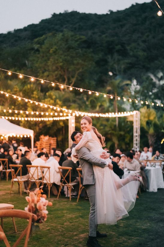Groom lifting bride at outdoor wedding reception. String lights and guests in background, mountain view.