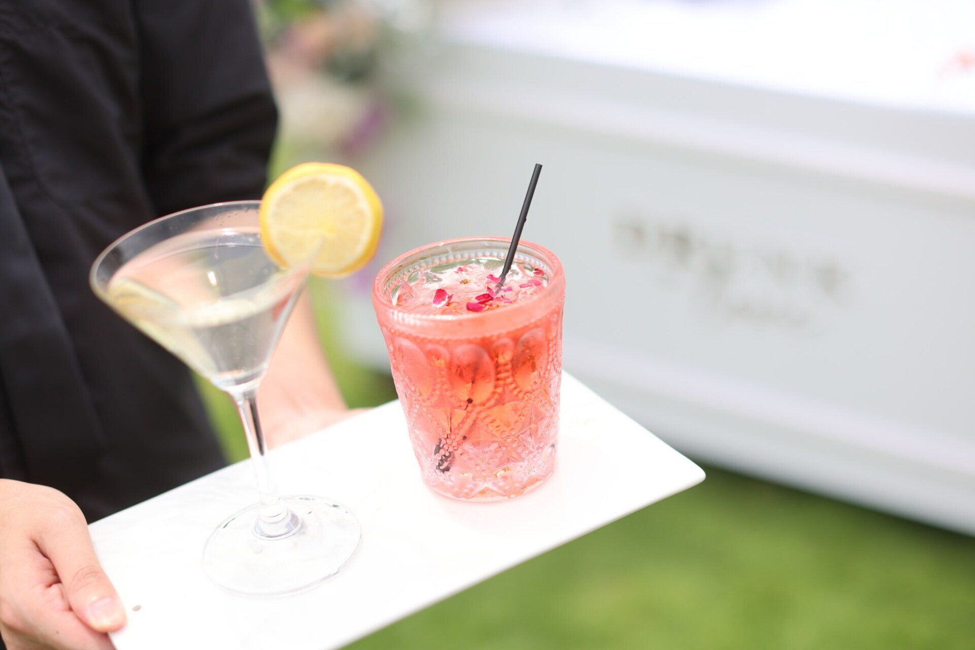 Person holding a tray with two cocktails: a clear martini with lemon and a pink drink with a straw.