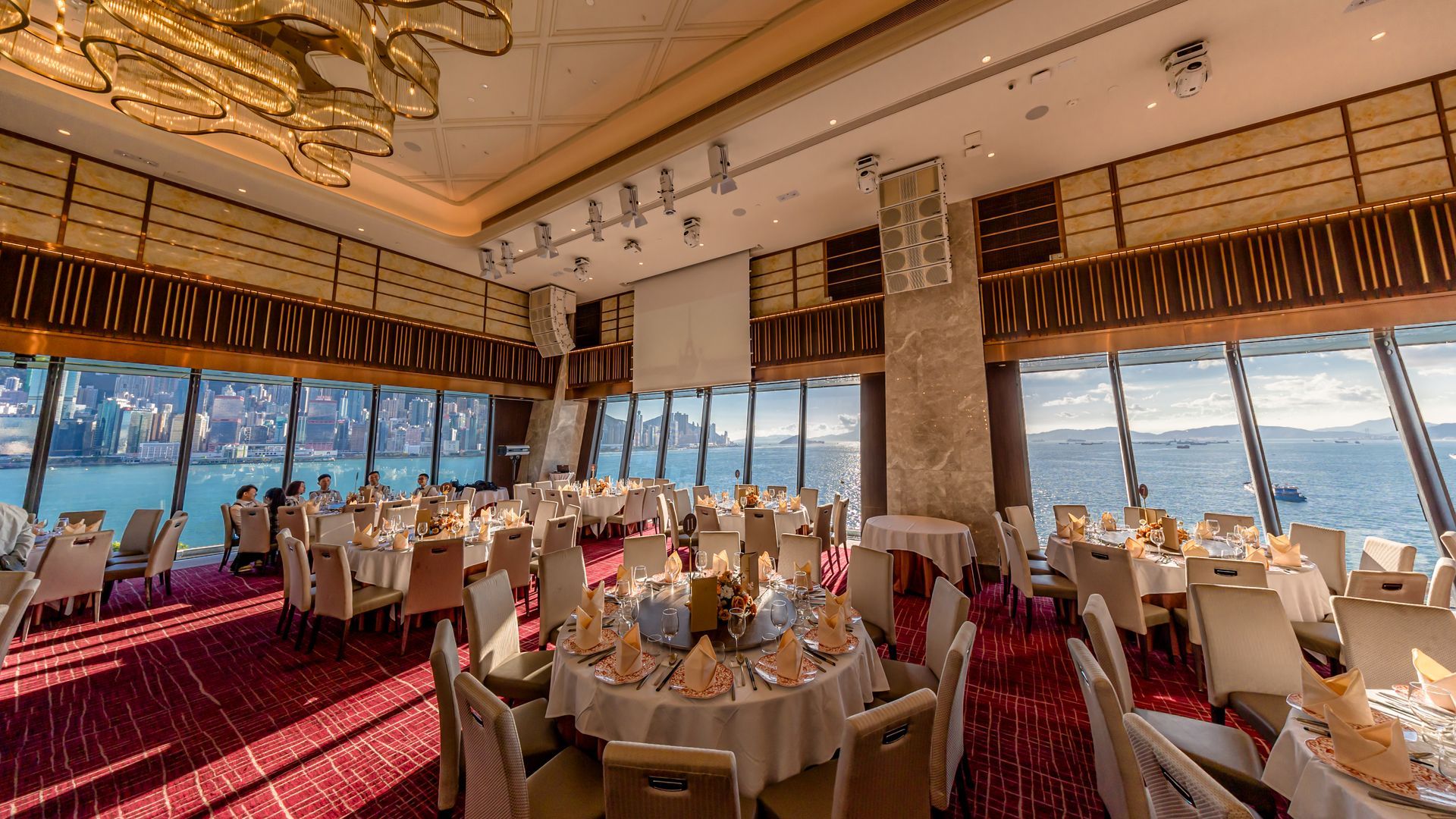 Banquet hall with round tables, large windows overlooking a city and water. Red carpet, cream chairs.