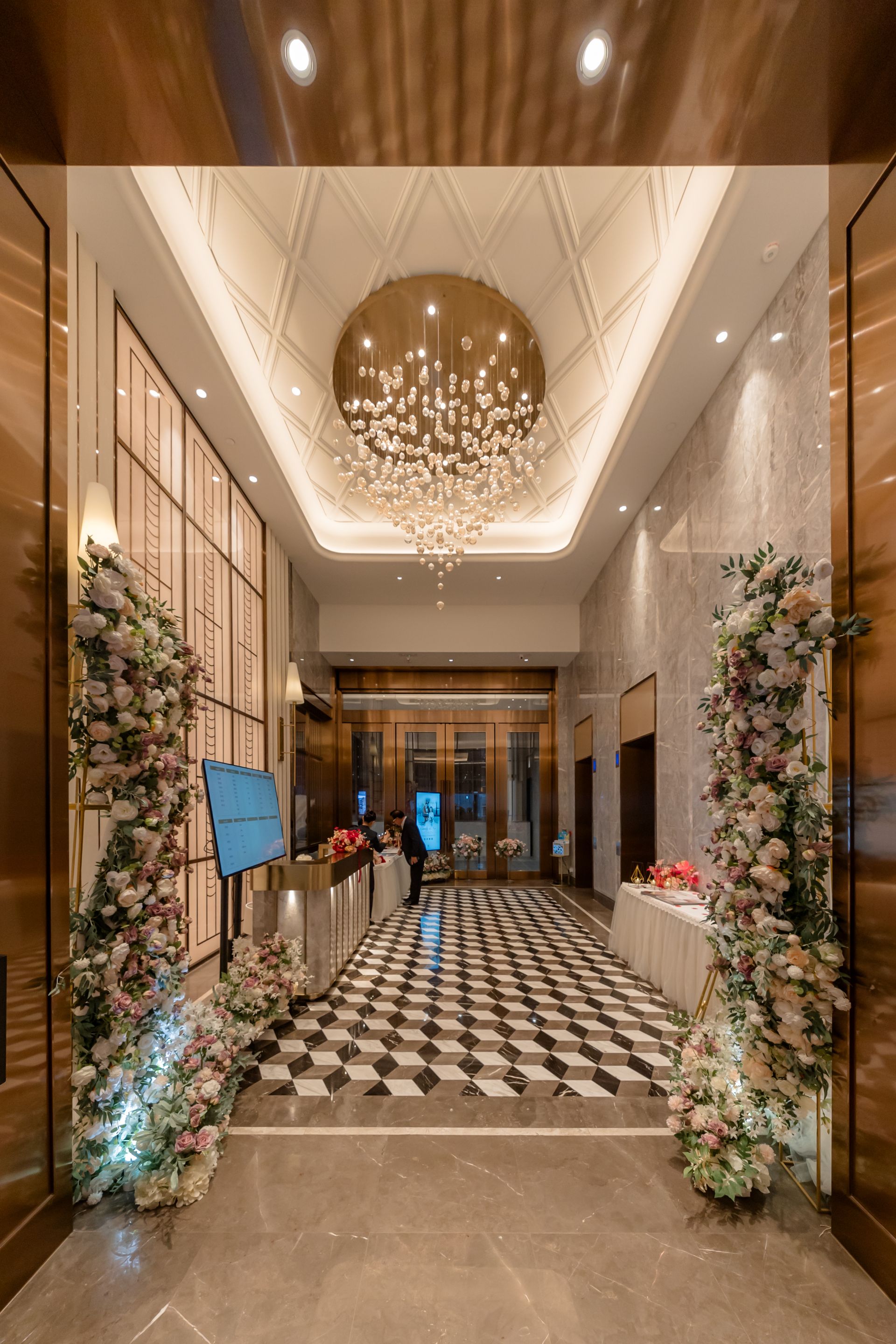 Elegant entrance with floral arrangements, checkered floor, chandelier, and a long hallway.