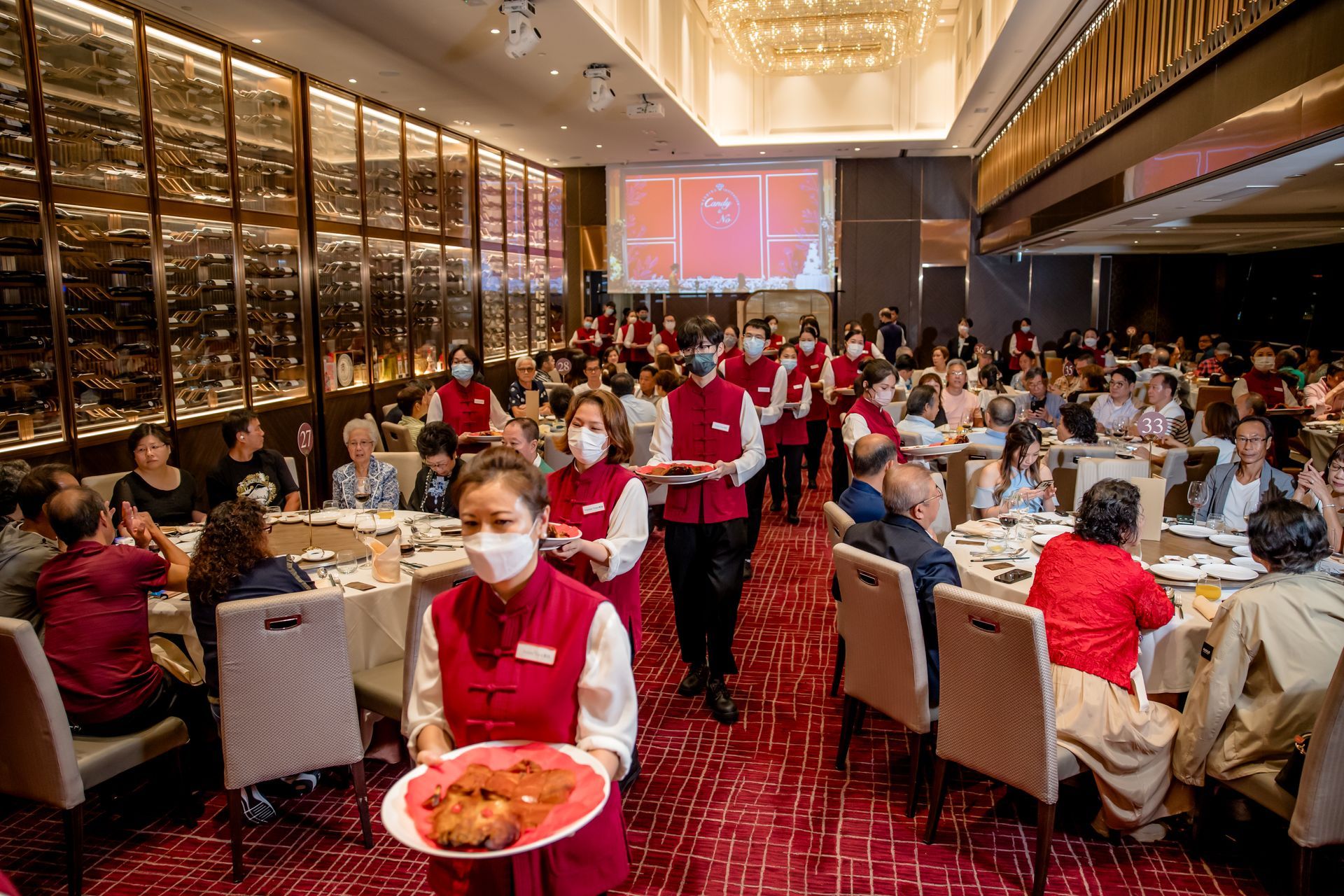 Waitstaff serving food in a crowded dining room with red carpet and wine display. Many seated guests.