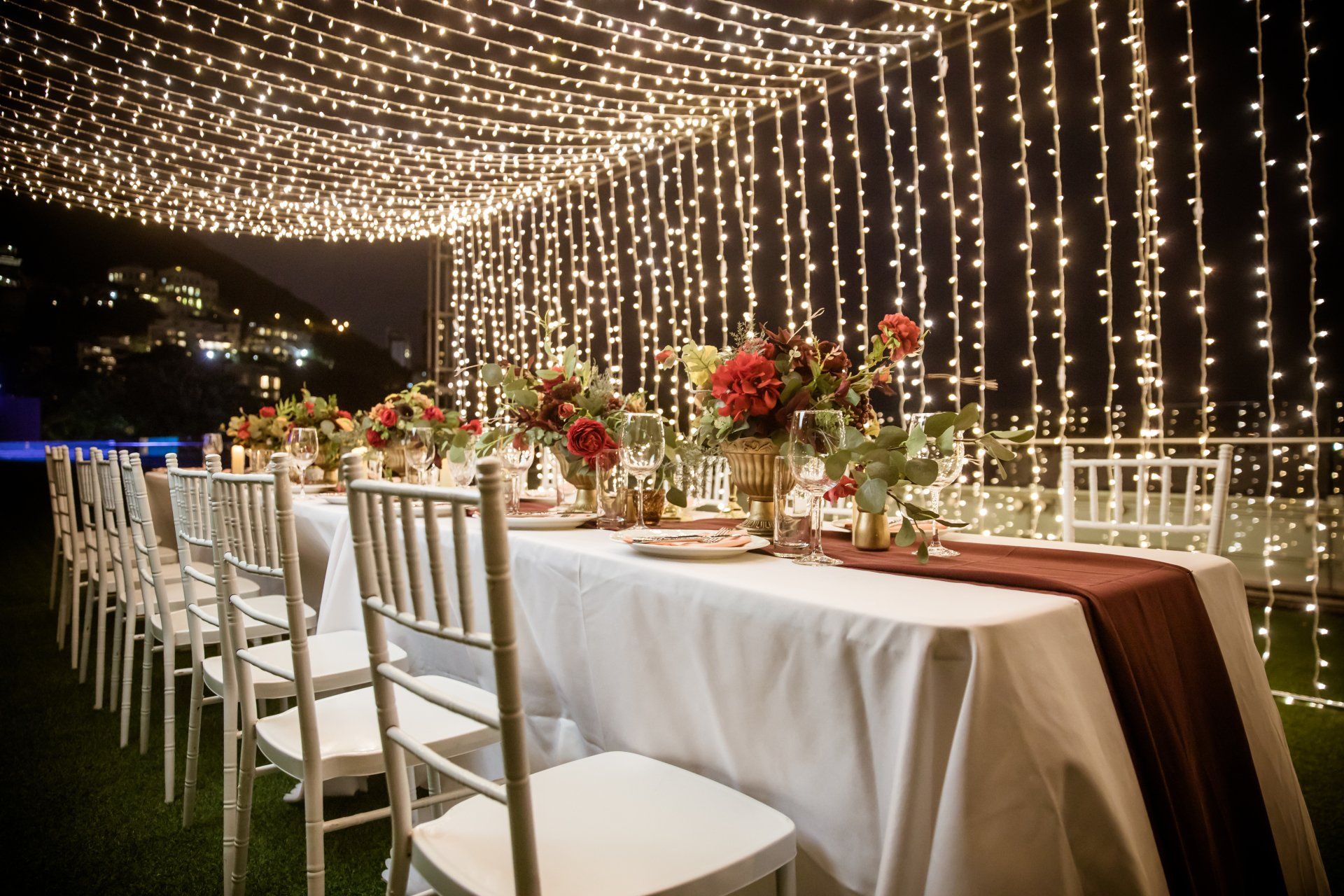 Long table set for a dinner outdoors, lit by strings of lights. Burgundy floral centerpieces and white chairs.
