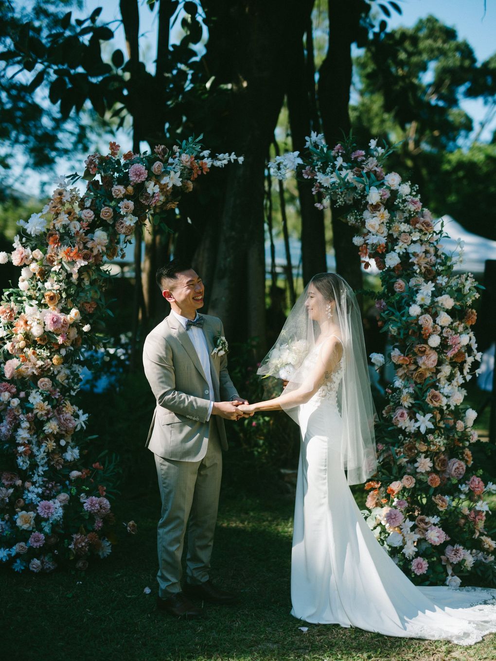 Couple holding hands under floral arch during an outdoor wedding ceremony.