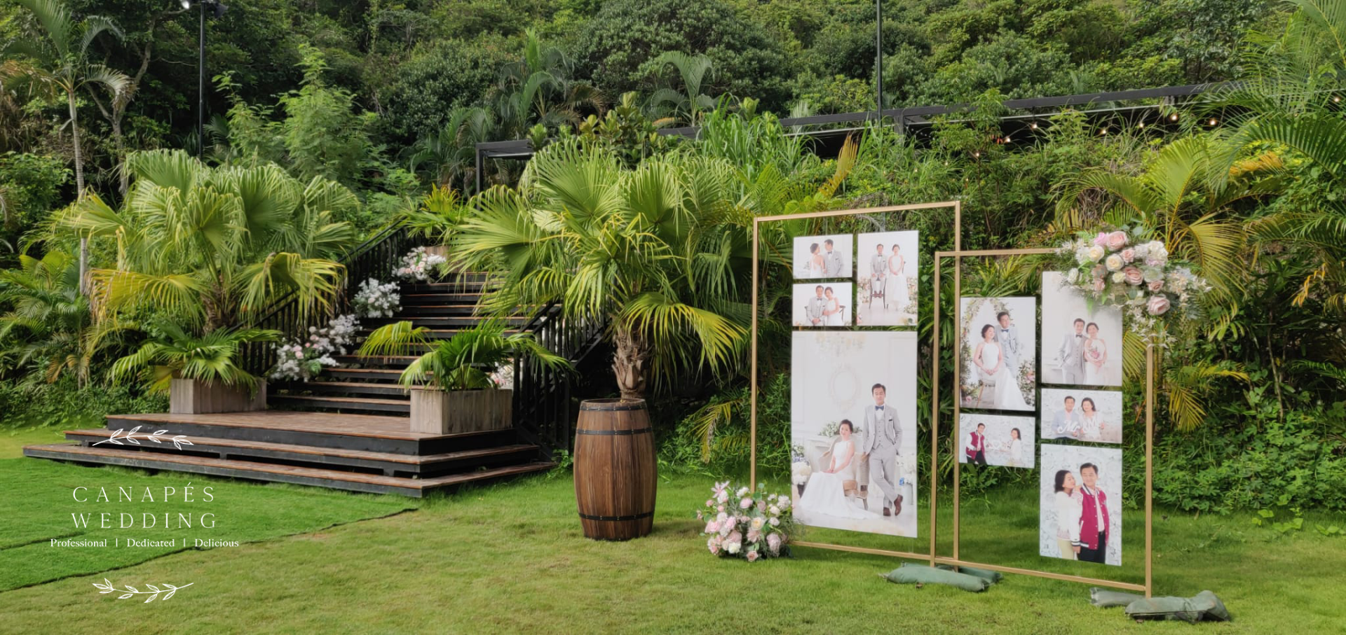 Wedding decor: photo displays, a stage, and a wooden barrel in a lush, green outdoor setting.