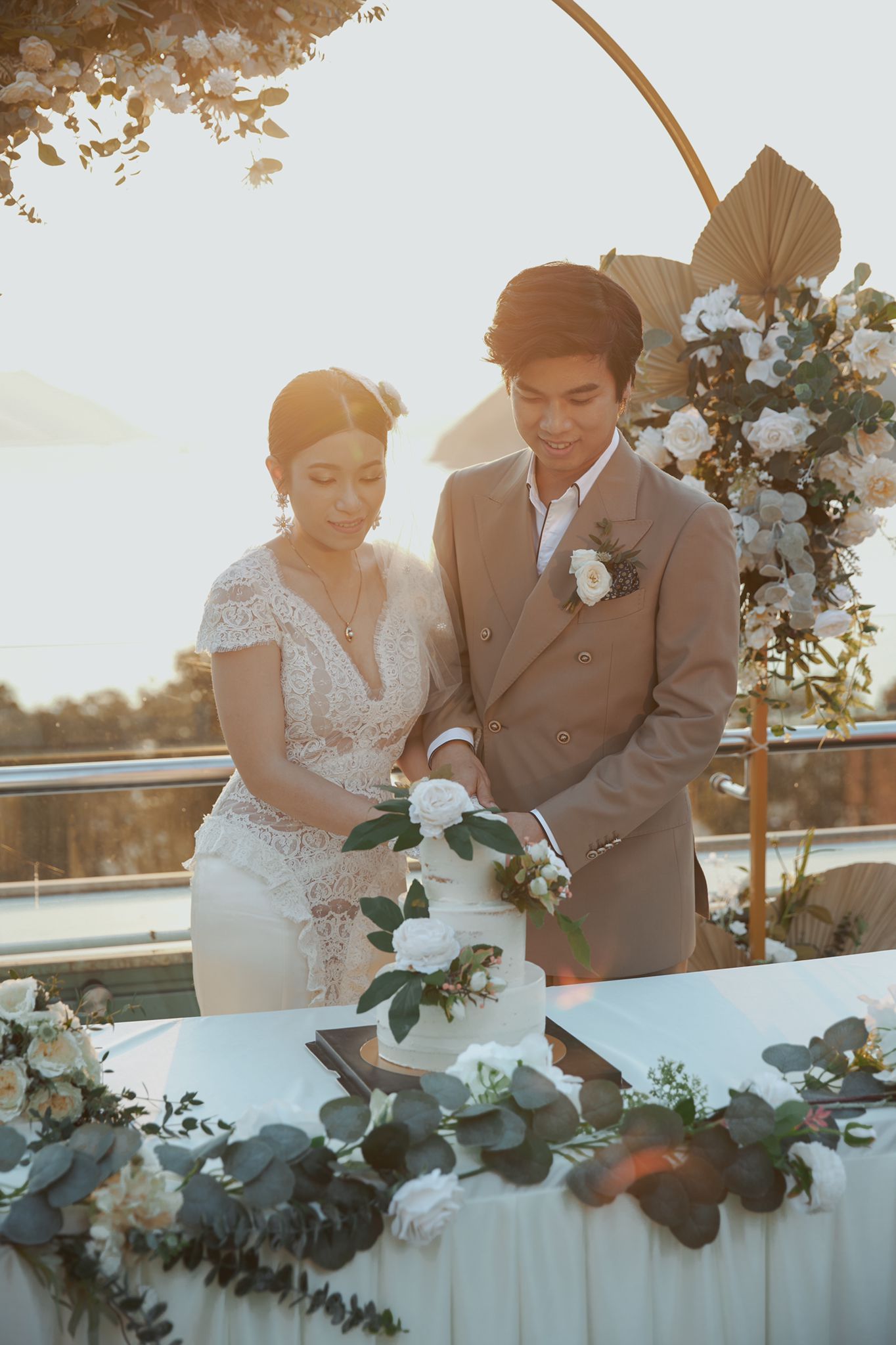 Couple cutting a white wedding cake outdoors, sunny backdrop, with floral decorations.