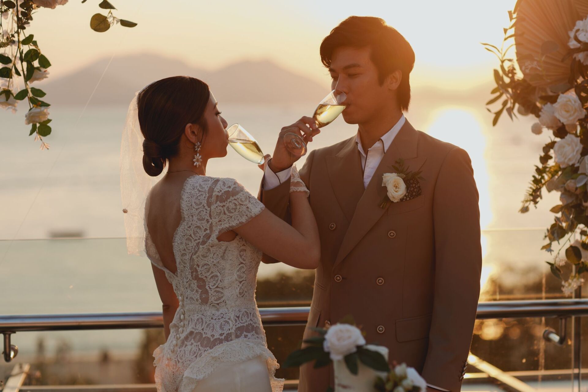 Couple toasting champagne, ocean sunset background, floral arch. Bride in lace gown, groom in suit.