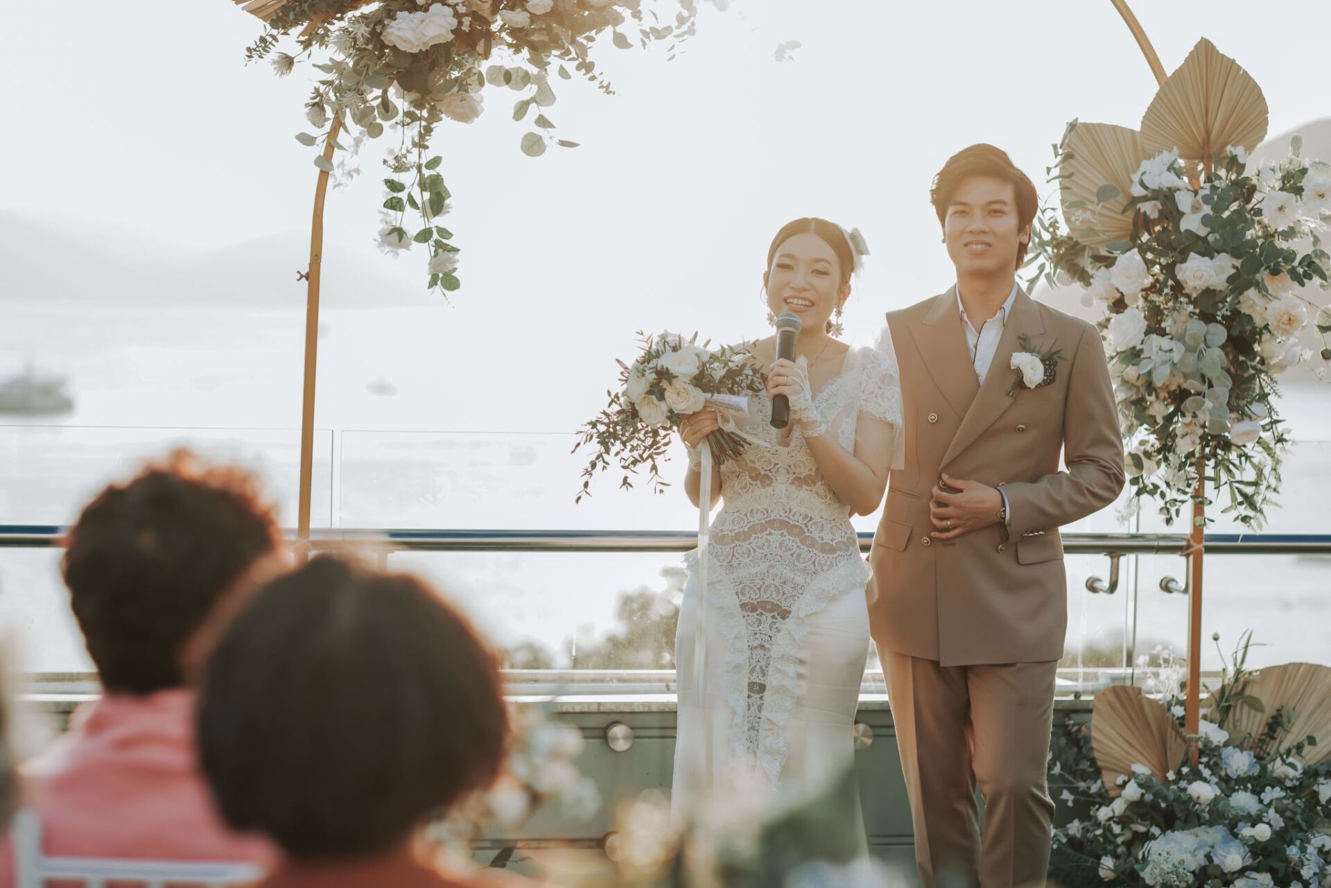 Two people at an outdoor wedding ceremony, one in a white dress, the other in a brown suit.