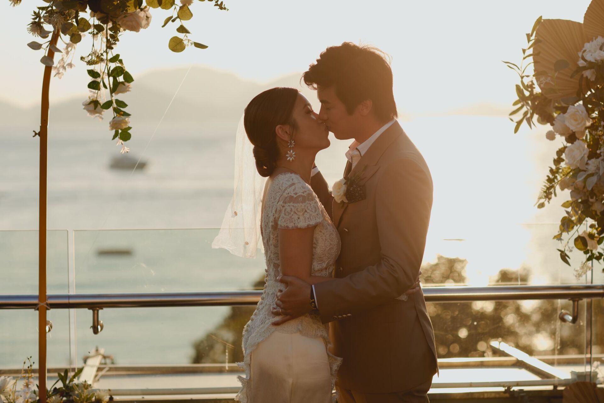 Couple kissing at outdoor wedding ceremony; sunset over water in background.