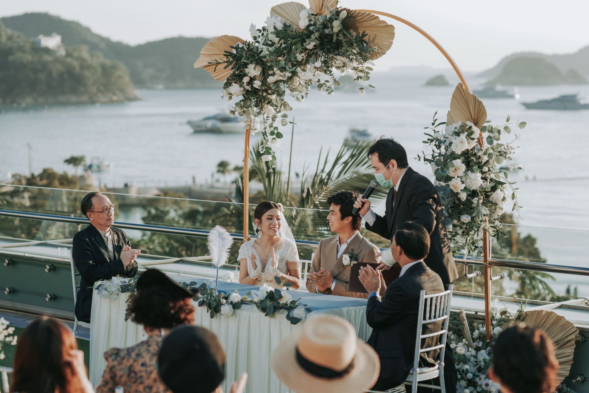 Wedding ceremony with couple at a table, floral arch, officiant speaking, ocean background, guests clapping.