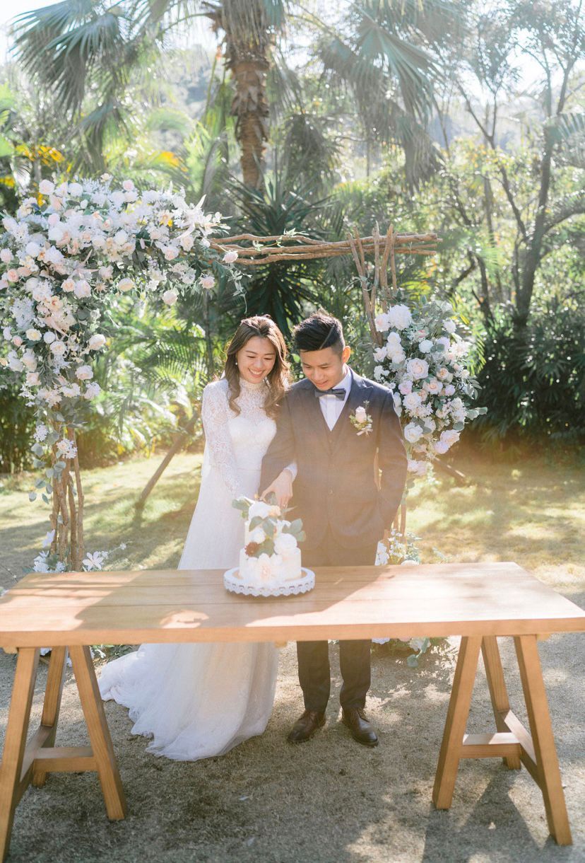 Couple cutting a wedding cake outdoors with floral arch backdrop.