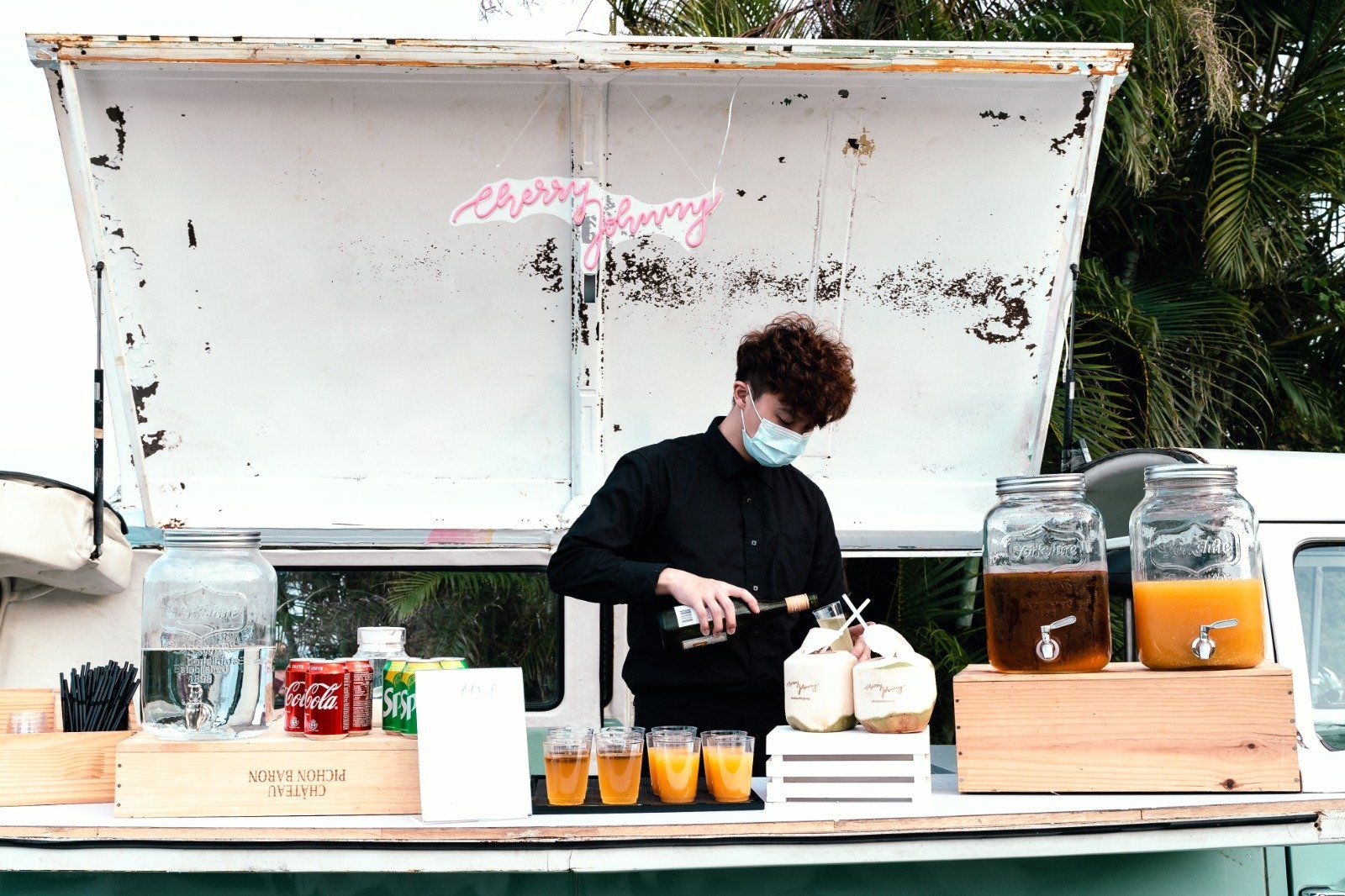 Person in mask at beverage stand, pouring drink. Beverages and coconuts on counter. White van.