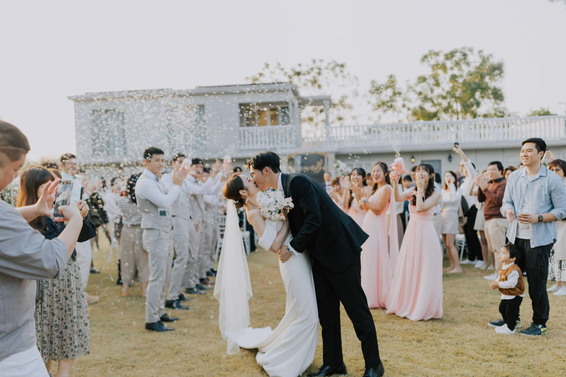 Newlyweds kiss as guests cheer, throwing confetti. Outdoor setting with a white building.