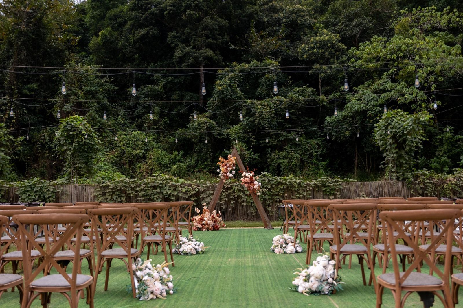 Wedding ceremony setup with wooden chairs facing a floral arch and a green lawn, surrounded by trees.